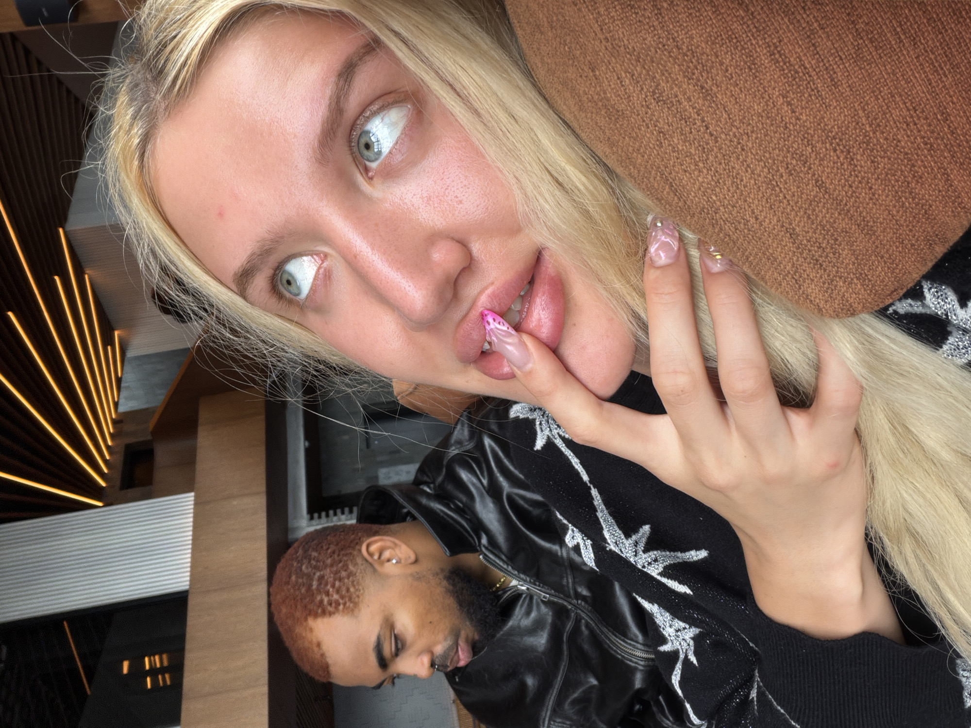 A close-up, selfie-style shot shows a light-skinned woman with blonde hair, looking upwards, with her finger touching her lips. Her nails are decorated with a butterfly design and gold accents. Beside her, a darker-skinned man with short, reddish-brown hair and a beard is visible in the background, wearing a black leather jacket. The environment appears to be indoors, with wooden paneling and ceiling lights that have an orange glow. There is no indication that this scene is located in Kintamani, Indonesia.