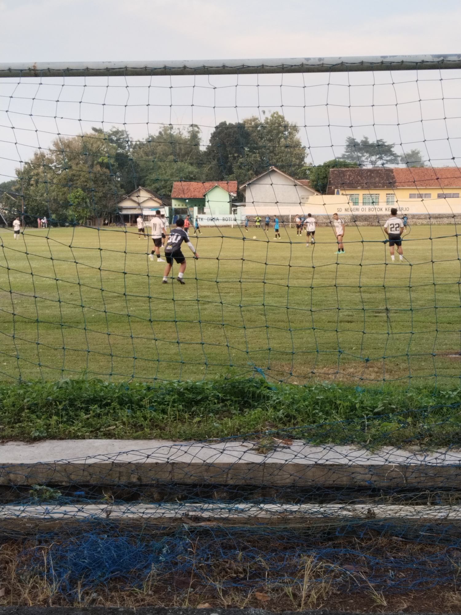 The image depicts a soccer game in progress at the "SD Negeri 01 Botok Kerjo" in Botok, Indonesia. Several players are visible on the grassy field, wearing numbered jerseys. A soccer net obscures the view, with overgrown vegetation and concrete structures in the foreground. Houses and trees are visible in the background. The sky is overcast.