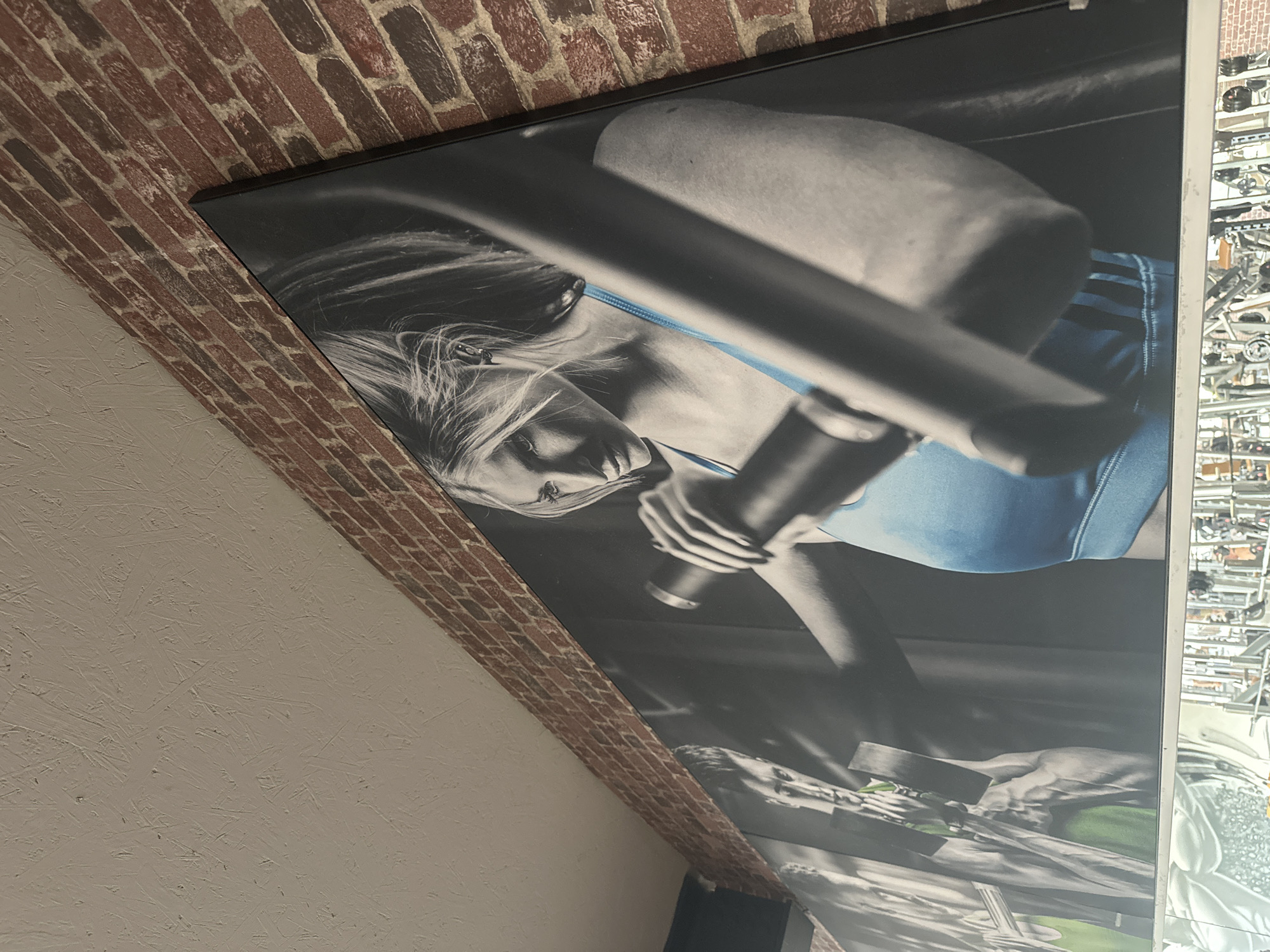 A vertically oriented photograph shows a framed picture hanging on a wall with exposed brick above and textured plaster below. The picture depicts a woman in athletic attire, likely exercising or participating in a fitness-related activity, based on the presence of gym equipment visible on the right side of the picture. The woman has blonde hair. The colors in the image are subdued, with black, white and blue and green, suggesting it may be a black and white photo that has been slightly colorized.
The specific location in Brunnthal, Germany cannot be determined from this photo.