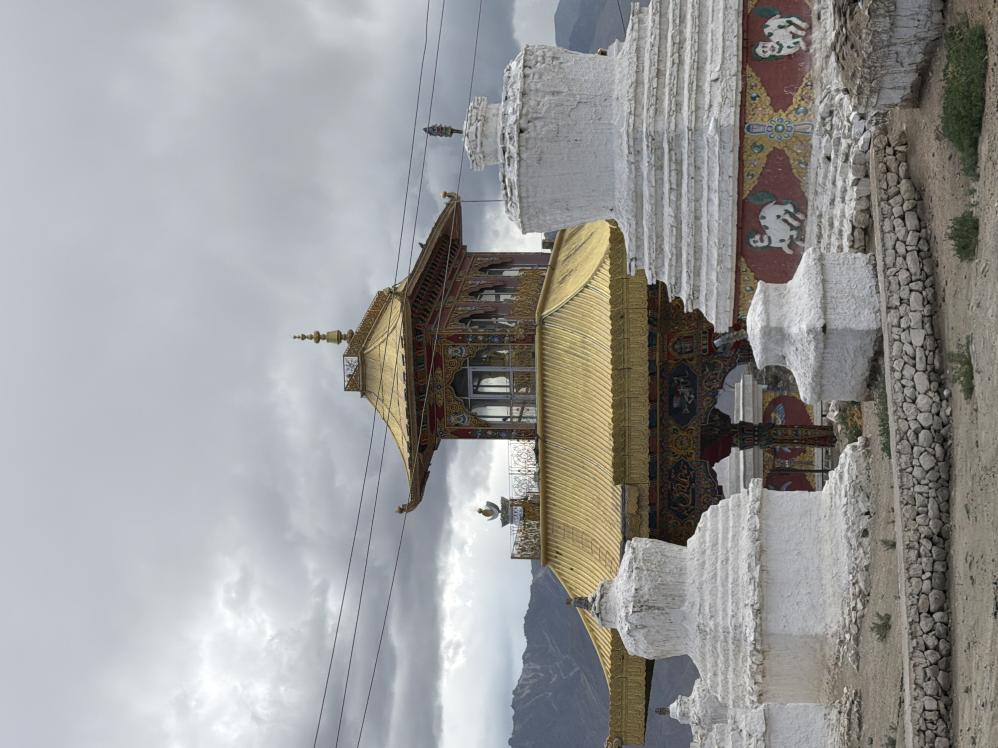This is a vertical, eye-level shot of the Leh Palace in Leh, India, under a cloudy sky. The palace, a prominent landmark, features a multi-tiered golden roof with intricate red and gold detailing on its eaves. Below the golden roof, there is a structure with arched windows and decorative elements. In the foreground, several white stupas of varying sizes and designs are present. The stupas are cylindrical and tiered, with some showing signs of age and wear. The larger stupas in the foreground have decorative bands with colorful motifs, including depictions of animals that appear to be dogs or similar creatures. The ground is a mixture of dry earth and gravel, with a low stone wall constructed in front of the stupas. In the background, a mountain range is visible, its peaks shrouded by the overcast sky. Power lines are strung across the upper part of the image, adding a modern element to the ancient setting. There are no people visible in the image.