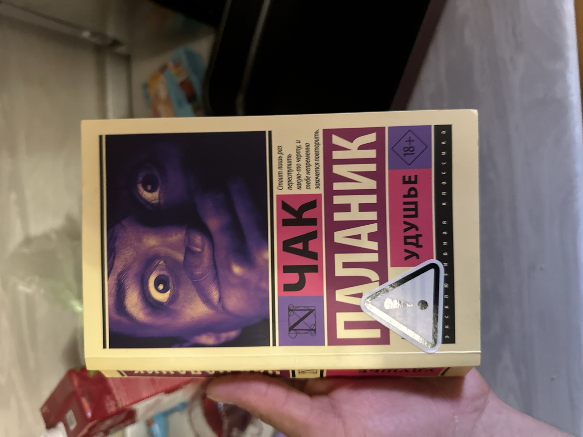 The image shows a close-up of a book cover held by a person's hand. The book has a striking design with a close-up of a person's face, eyes wide and mouth covered by their hand, rendered in purple and yellow tones. The title of the book appears to be "ЧАК ПАЛАНИК" (CHAK PALANIK), with the word "УДУШЬЕ" (UDUSH'YE) beneath it. There is a triangular warning symbol with an exclamation mark and an "18+" age rating symbol on the lower part of the cover. The background is blurred, showing what appears to be a kitchen or pantry area with some packaged goods. There is no information in the image to suggest any connection to Saransk, Russia.