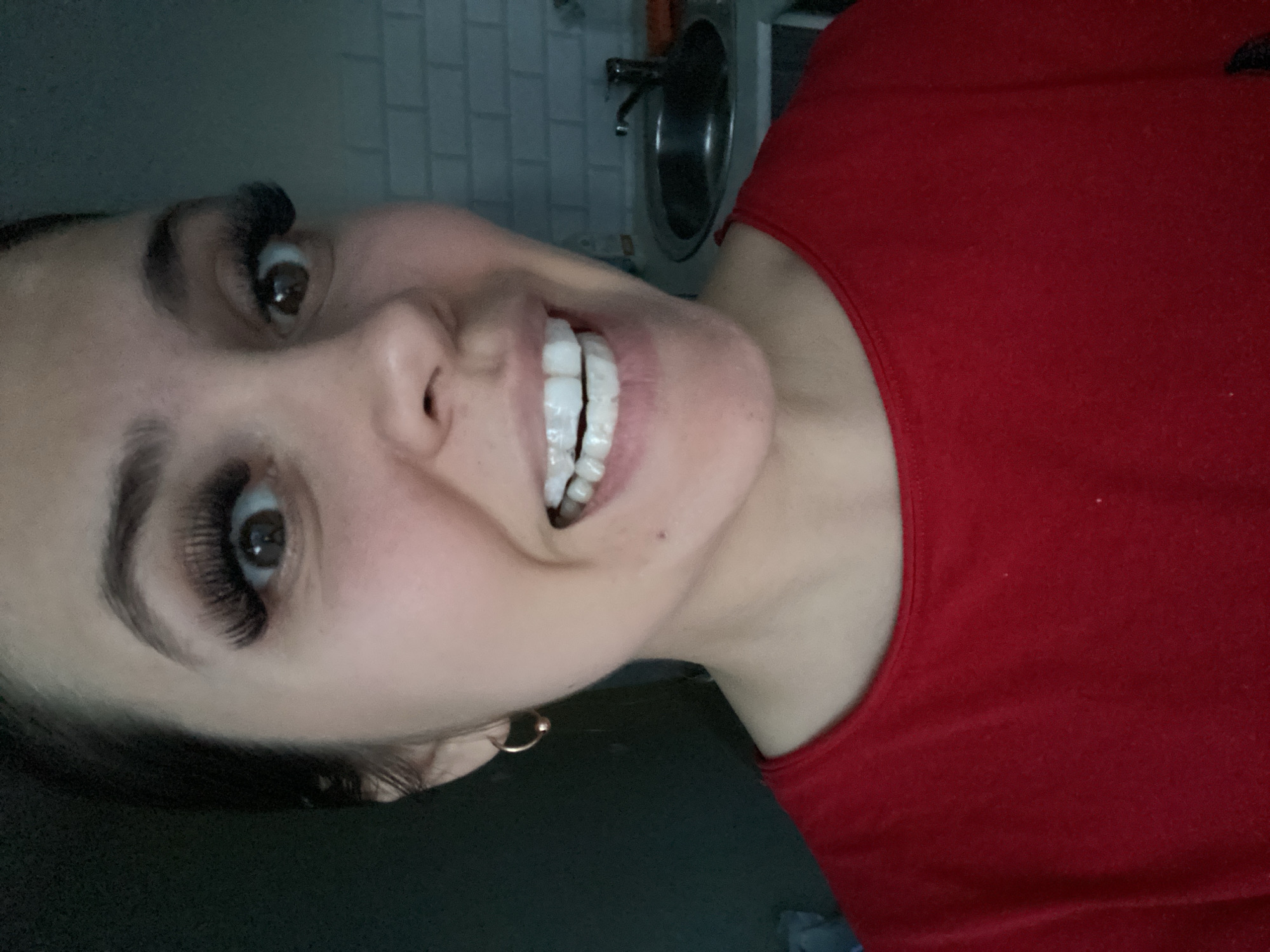 A close-up, rotated portrait captures a person, likely female, smiling broadly indoors. The image is oriented with the person's head towards the left side of the frame. The subject has dark hair, dark eyes, prominent, full eyelashes, and well-defined eyebrows. She is smiling widely, revealing a full set of very white, uniformly shaped teeth, suggesting veneers or dental work. A small, round, gold-colored earring is visible in her left earlobe. She is wearing a simple red top, possibly a tank top or t-shirt, which covers her shoulder and part of her chest.
The background shows an indoor setting, likely a kitchen or bathroom. Visible elements include light-colored, possibly white or grey, tiled walls (resembling subway tiles) and a stainless steel sink with a metallic faucet. The lighting is somewhat dim, casting subtle shadows, and appears to be artificial indoor light. The expression on the person's face conveys happiness or amusement. There is no visible text in the image.