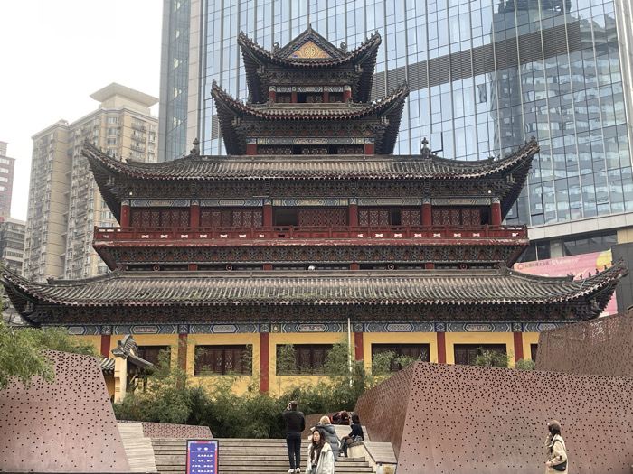 An urban scene in Yuzhong District, China, features a prominent, multi-tiered traditional Chinese temple structure centrally positioned. The temple exhibits dark, ornate curved roofs, deep red walls on its upper levels, and a yellow base on its lowest tier, all adorned with intricate colorful patterns and lattice work. Its traditional architecture directly contrasts with the modern surroundings: a tall, reflective glass skyscraper rises immediately behind the temple on the right, and a lighter-colored residential high-rise stands in the background to the left.
In the foreground, a wide set of light-toned steps ascends towards the temple's entrance. Flanking these steps are two angled, reddish-brown retaining walls or decorative panels, each featuring a pattern of numerous small, evenly spaced circular perforations. Green foliage, resembling bamboo, grows behind these perforated panels and at the base of the temple.
Several individuals are present: a group of approximately three to four people are walking up the steps towards the temple, one wearing a dark jacket. Another individual in a light-colored coat and hat is visible in the lower right foreground, moving away from the viewer. The sky is overcast and grey, indicating diffused lighting conditions.