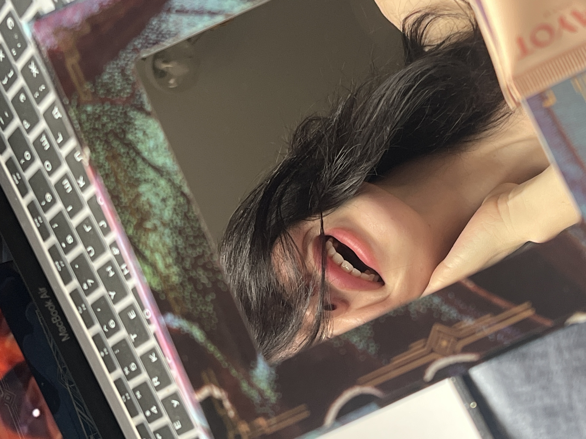 Here's a detailed description of the image:
The image shows a reflection in a mirror. The reflection includes a person's face, with dark hair, visible teeth, and a hand holding the mirror. The face is partially obscured by strands of hair. The mirror is resting on a desk, next to a MacBook Air. A tube of "AYOT" lotion is visible in the upper right corner. The mirror has a decorative frame with a mottled, dark green and brown pattern. The person appears to be indoors, and the overall environment is somewhat cluttered. There is no information in the image to identify the location as Krasnodar, Russia.