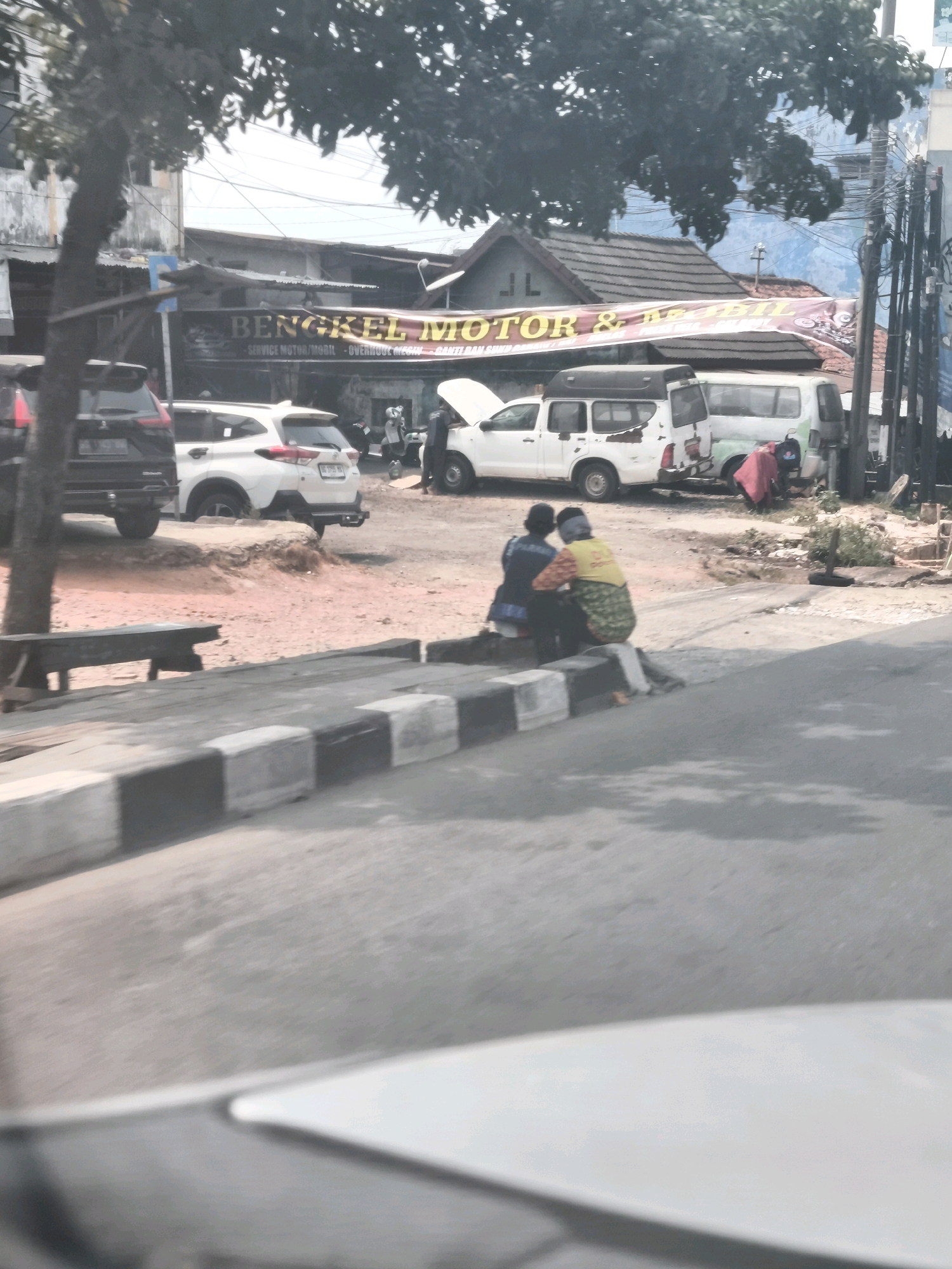In Palembang, Indonesia, two individuals are seated on a curb adjacent to a street. The person on the left wears a dark blue shirt and dark pants, with their head covered by a dark headscarf. The person on the right is dressed in a yellow shirt with a green and white patterned garment draped over their lap and shoulders. They are positioned in front of a row of vehicles, including a white pickup truck with an open hood and a white van. Behind the vehicles, a banner is displayed with text that appears to read "BENGKEL MOTOR & MOBIL" which translates to "Motorcycle and Car Workshop." The surrounding area shows buildings with tiled roofs, a tree, and a street with a concrete barrier.
