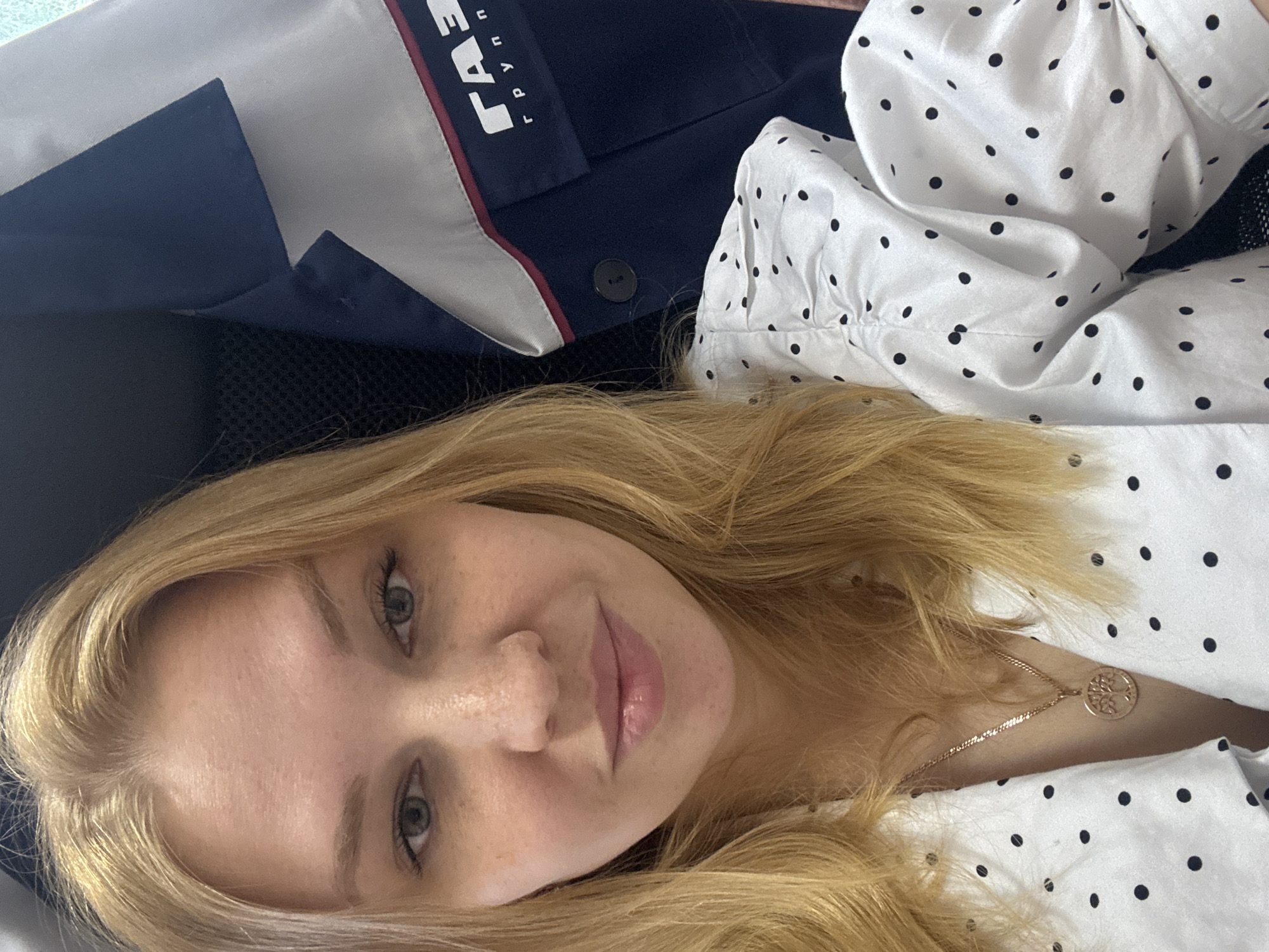 A young woman with blonde hair and fair skin is captured in a close-up, slightly angled selfie. She is wearing a white blouse with black polka dots and a necklace with a circular pendant. Behind her, a navy blue jacket with white and red trim is visible, featuring a logo that reads "ГАЗ групп". The background appears to be an indoor setting, possibly an office or vehicle. The overall impression is a casual, personal photograph. The location is identified as Zavolzhye, Russia.