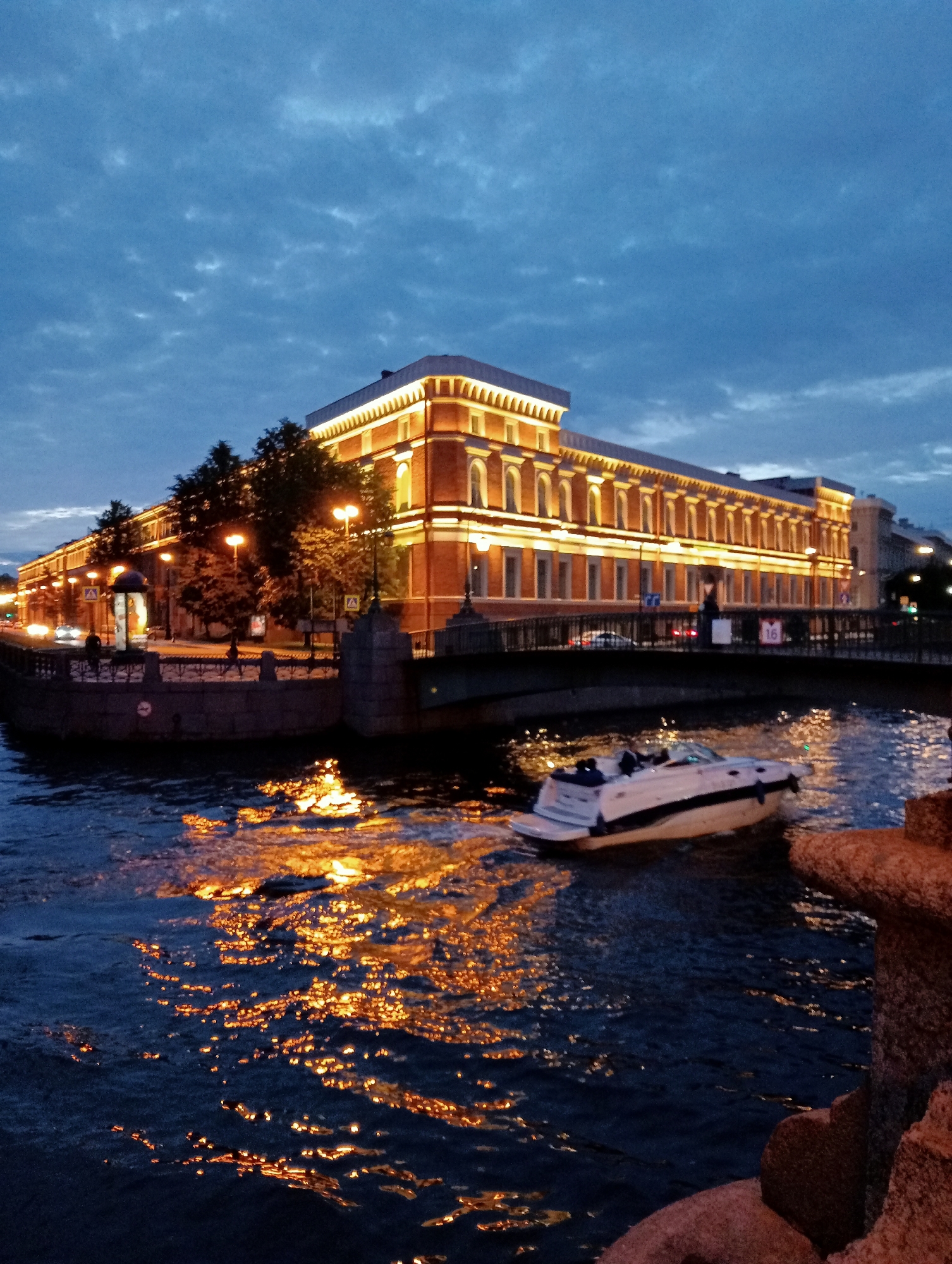 This is a photograph of the St. Petersburg Canal at dusk. The scene features a canal with a motorboat in the foreground, moving from right to left. The boat is white with black trim and has at least two people on board. The water reflects the warm, golden light from the streetlights and illuminated windows of the buildings lining the canal.
On the left side of the canal, there is a multi-story brick building with a modern facade, brightly lit by numerous yellow lights, highlighting architectural details such as arched windows and decorative cornices. Trees with dense foliage are visible between the building and the canal. A pedestrian walkway with a dark railing runs along the edge of the canal. Streetlights are spaced along this walkway. A bridge, also lit, spans the canal in the background, with the faint headlights of cars visible on it.
The sky is a deep blue with scattered, wispy clouds, indicating either twilight or pre-dawn. On the right side of the frame, a rough, textured stone structure, possibly part of a bridge or embankment, is visible in the foreground, adding depth to the composition. The overall atmosphere is serene, with the artificial lights creating a vibrant contrast against the darkening sky and water. The location is likely near the center of St. Petersburg, given the architectural style and the presence of canals and illuminated historic buildings.