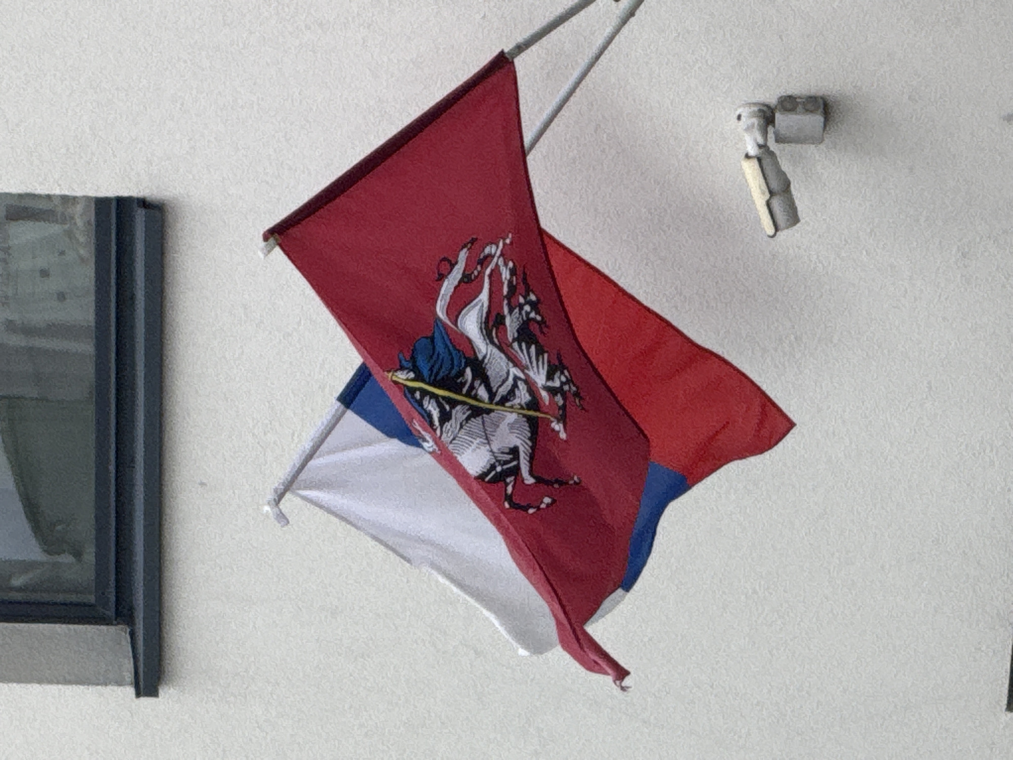 Two flags, a Russian flag and a red flag with a white equestrian image, are displayed on a textured white wall. The Russian flag, partially visible, has white, blue, and red horizontal stripes. The red flag features a detailed depiction of a knight on a horse, a symbol of Saint George slaying a dragon, which is the coat of arms of Moscow. Both flags are attached to poles, one of which is metallic and extends diagonally upwards. A security camera is mounted on the wall to the right of the flags. Above the flags, a window with dark framing is visible against the wall. The scene appears to be outdoors, with the flags waving gently.