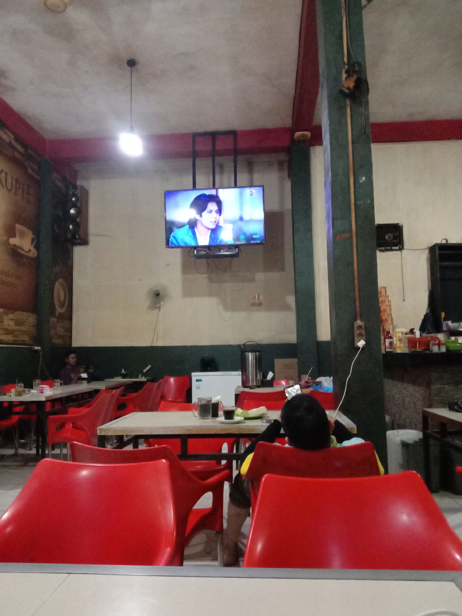 This image depicts the interior of a casual cafe or eatery in Lhokseumawe, Indonesia, likely in the evening given the reliance on artificial lighting. The space features light-colored walls with a darker green base trim, concrete ceilings, and prominent dark green support columns. A very bright, single light bulb hangs from the ceiling, illuminating the central area.
In the foreground, a person with dark hair, wearing a yellow-sleeved shirt, is seated in a red plastic chair, facing away from the camera. They are holding a smartphone and appear to be watching a flat-screen television mounted high on the wall, which shows a person with dark hair wearing a blue jacket. Another person is dimly visible seated at a table on the far left.
The cafe is furnished with numerous red plastic chairs and simple tables with light-colored tops and dark metal frames. Several tables hold glasses of beverages (including water and coffee) and small plates. A large graphic on the left wall displays the partially visible text "KUPIE," likely referring to coffee. Other objects include a speaker mounted near the TV, a white refrigerator with "BSA" visible on its side, a thermos, and various supplies behind a counter in the background. Electrical outlets and wiring are visible on a support column. The overall atmosphere is relaxed and informal.