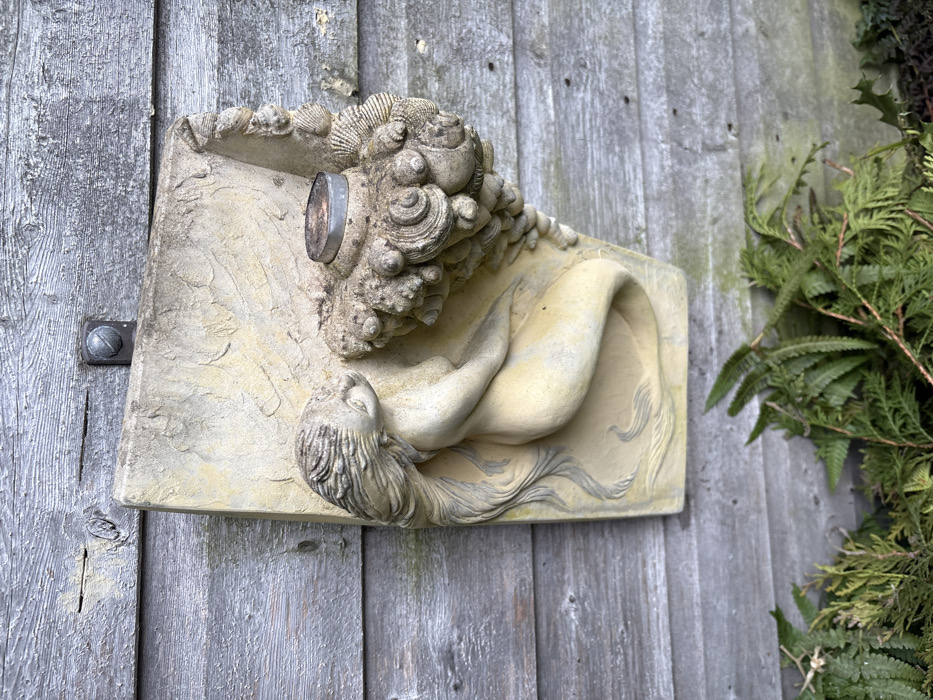 This image displays a weathered, light-colored decorative plaque affixed to a grey, horizontally planked wooden wall in Stiperstones, United Kingdom. The plaque depicts a mermaid-like figure with long, flowing hair and a curved tail, reclining towards the left. To the right of the figure, an ornate, textured mass resembling coral and seashells features a recessed cylindrical metal object, possibly a candle holder or sconce, at its apex. The plaque is secured to the wall by a single metal bolt and washer visible at the top center. The wooden wall exhibits aging and wear. At the base of the image, green foliage, primarily ferns and conifer branches, is visible on the ground level. No individuals are present in the scene.