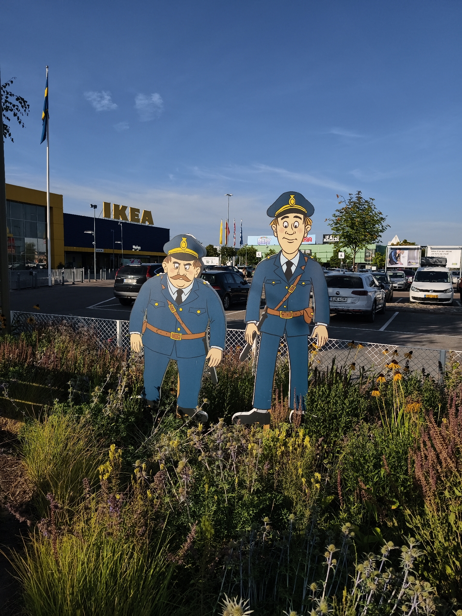 The image captures a scene outside an IKEA store in Linköping, Sweden. Two life-size cardboard cutouts of police officers stand in the foreground, positioned in front of a flowerbed. One cutout depicts a heavier-set officer with a mustache, wearing a blue uniform, a dark cap with a gold emblem, and a belt with a holster. The other cutout is of a slimmer officer, also in a blue uniform and cap, with a simpler facial expression.
Behind the flowerbed and the cutouts is a parking lot filled with cars. In the background, the distinctive blue and yellow architecture of the IKEA store is visible, with the "IKEA" logo prominently displayed. A Swedish flag flies from a tall flagpole to the left of the building. The sky is clear and blue with a few wispy clouds, and the sun casts shadows, indicating it is likely daytime. The overall environment suggests a typical retail store exterior on a bright day.