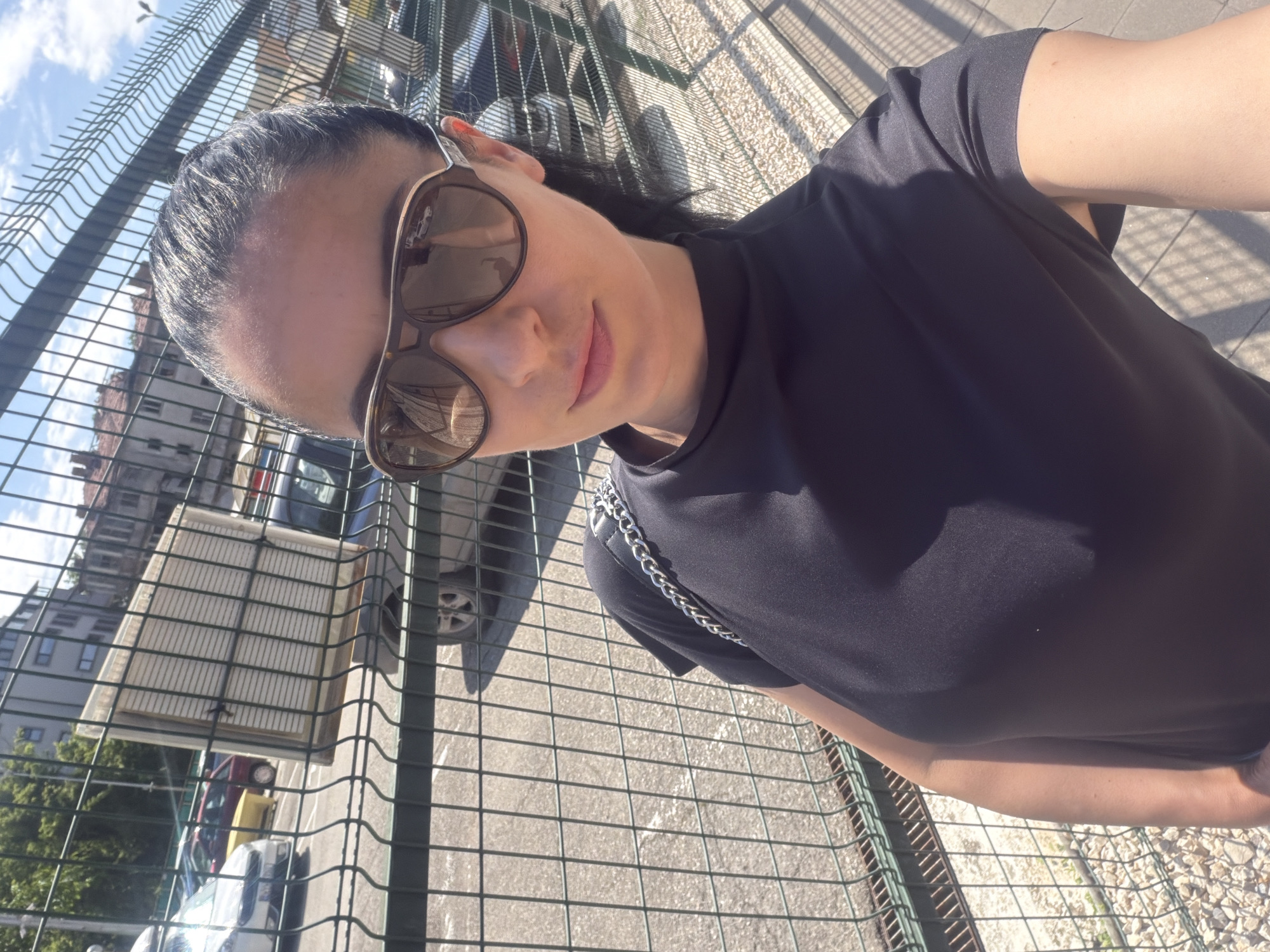 A woman is captured in a sunny outdoor selfie, positioned in front of a prominent green chain-link fence. She has dark hair, pulled back, and is wearing large, dark-framed sunglasses that reflect the bright sky and parts of the surroundings. Her expression shows a faint, pleasant smile. She is dressed in a plain black t-shirt, and a thin silver chain with large links is visible draped over her left shoulder and chest area.
The background, seen through and beyond the fence, indicates an urban setting in Pleven, Bulgaria. Behind the fence, there are various buildings, some with multiple windows, and several parked cars (including red, yellow, and white vehicles). Green foliage from trees is also visible. The ground appears to be a paved or gravel surface, with strong shadows cast by the fence and other elements, characteristic of a bright, sunny day, likely midday or early afternoon. The sky, visible above the fence, is clear with some scattered clouds. The image is taken at an angle, giving it a dynamic, slightly tilted perspective. There is no visible text in the image.