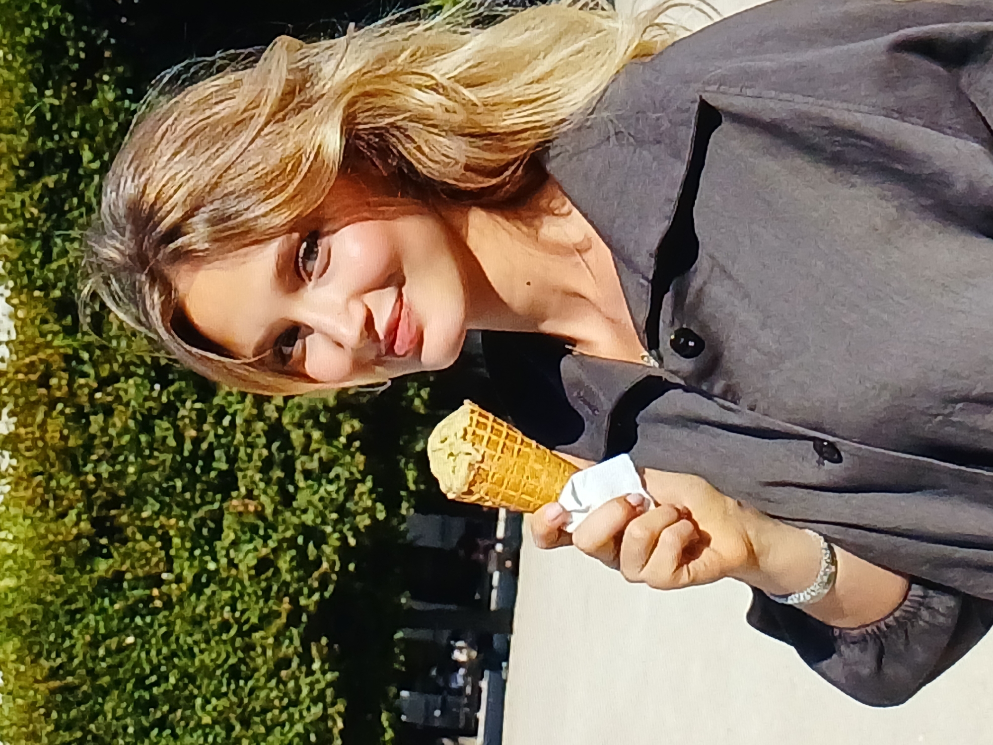 A young woman with blonde hair is holding an ice cream cone and looking at the camera. She is wearing a dark grey jacket. In the background, there are trees and a paved area. The image is taken outdoors during the day.