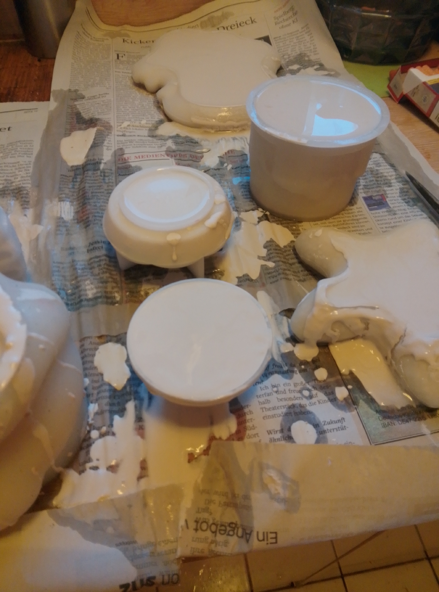A close-up, high-angle shot shows a surface covered with newspaper and white, liquid material that appears to be plaster or paint. The material is spread unevenly, with some areas appearing as dripped or poured puddles. Several round objects, possibly lids or containers, are partially submerged in the white substance. One of these objects is a white, cylindrical container with a clear plastic lid. Another is a flat, round white object with a textured bottom. The newspaper beneath is also wet and slightly torn in places. The overall scene suggests a crafting or DIY project in progress. There is no indication of the location being the city of Wedel, Germany.