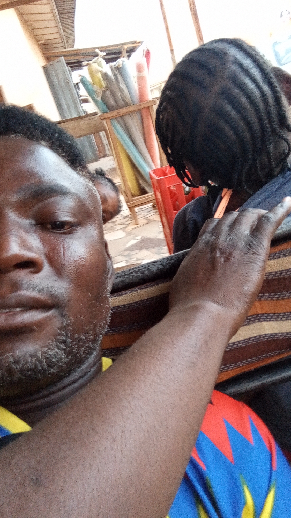 The image presents a close-up, angled view of a scene in what appears to be an outdoor or semi-outdoor setting, likely in Sabon Tasha, Nigeria. In the immediate foreground, a dark-skinned man is visible, occupying the left side of the frame. His face, partially obscured, shows a short, dark beard with some gray, and a neutral to slightly serious expression. His left arm extends across the frame, resting on a striped fabric, possibly a chair or bench. He wears a multi-colored shirt featuring blue, red, and yellow patterns.
Behind the man, towards the right, is another person seen from the back, characterized by dark, tightly braided hair. Further in the background, a faint and blurry outline of a child's head is visible. The background reveals details of a commercial environment, possibly a market stall or roadside shop. A wooden structure holds several rolled-up fabrics or mats in various colors including yellow, light blue, grey, and pink. A red plastic crate is also visible on the ground. Overhead, corrugated metal sheets suggest part of a makeshift roof structure. The ground is paved with what appear to be patterned tiles or bricks. The bright lighting indicates it is daytime. No discernible text is visible within the frame. The scene conveys a sense of everyday activity in a lively public space.