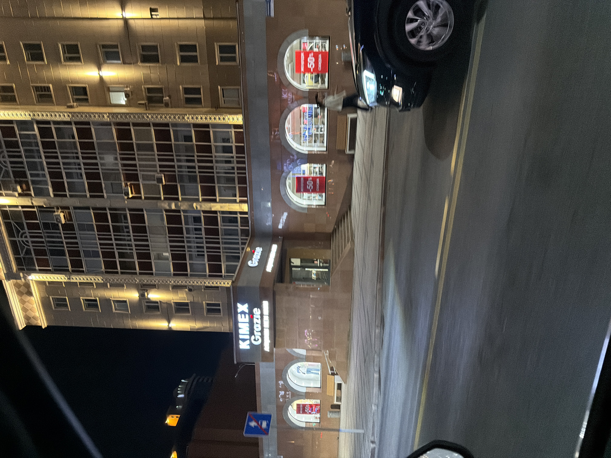 The image depicts a nighttime street view in Astana, Kazakhstan, focusing on the facade of a commercial building. The building features a prominent sign that reads "KIMEX Grazie" in white lettering. Below the sign, several arched windows display merchandise, with prominent red signs indicating "-50%" discounts. A person is visible walking past one of these windows. A black car is in motion in the foreground, its headlights illuminated. The street is paved, with yellow lane markings visible. The overall lighting suggests an urban environment at night, with artificial lights illuminating the building and street.
