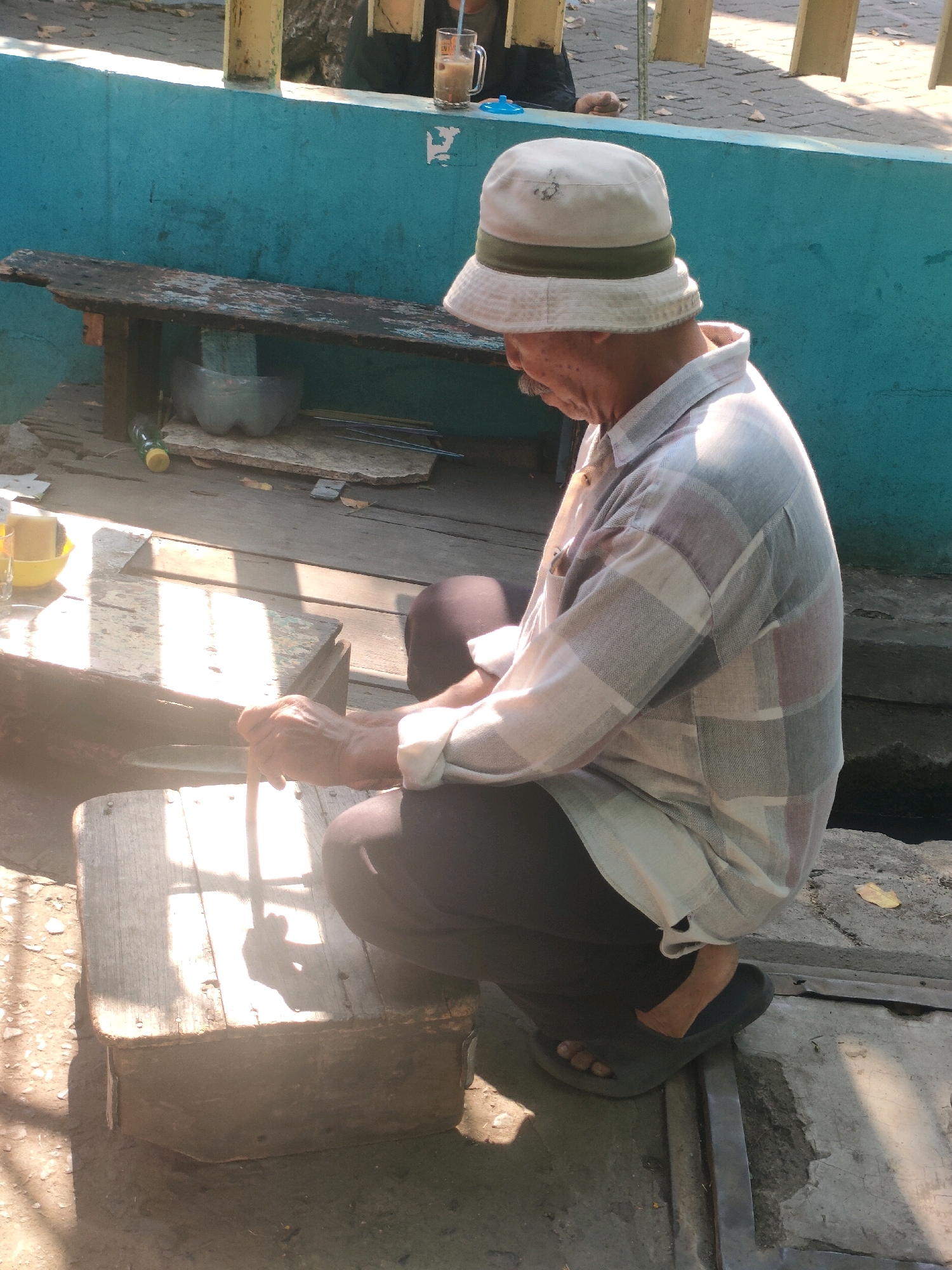 An elderly man, wearing a bucket hat and a plaid shirt, squats on a wooden structure next to a blue wall. Sunlight casts strong shadows across the scene, illuminating the wooden planks and the man's hands, which hold a cane. A glass of iced coffee sits on a bench in the background, and a plastic bottle is visible on the ground. The setting appears to be an outdoor area, possibly in Surabaya, Indonesia, with elements suggesting a casual or street-side environment.