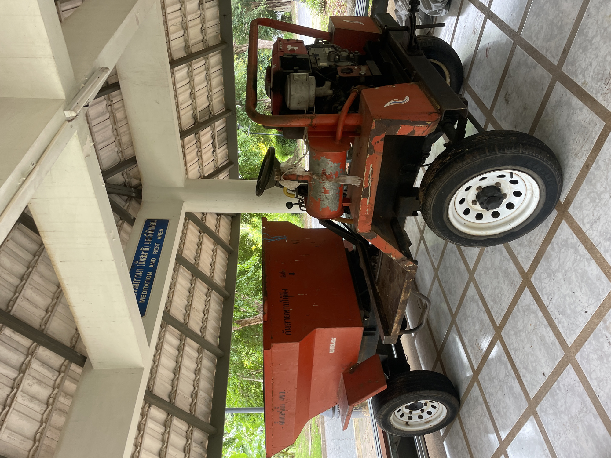 An orange, four-wheeled utility vehicle with a large hopper is parked indoors under a covered, tiled roof. The vehicle has a steering wheel, a small engine in the rear, and appears to be made of metal. White text is visible on the orange hopper, and a blue sign with white lettering hangs from the ceiling. The sign reads "MEDITATION AND REST AREA" in English and also has text in Thai. The floor is tiled with square, light-colored tiles. Outdoor greenery is visible through an open doorway in the background. The vehicle is likely in Na Chom Thian, Thailand, as indicated by the sign.