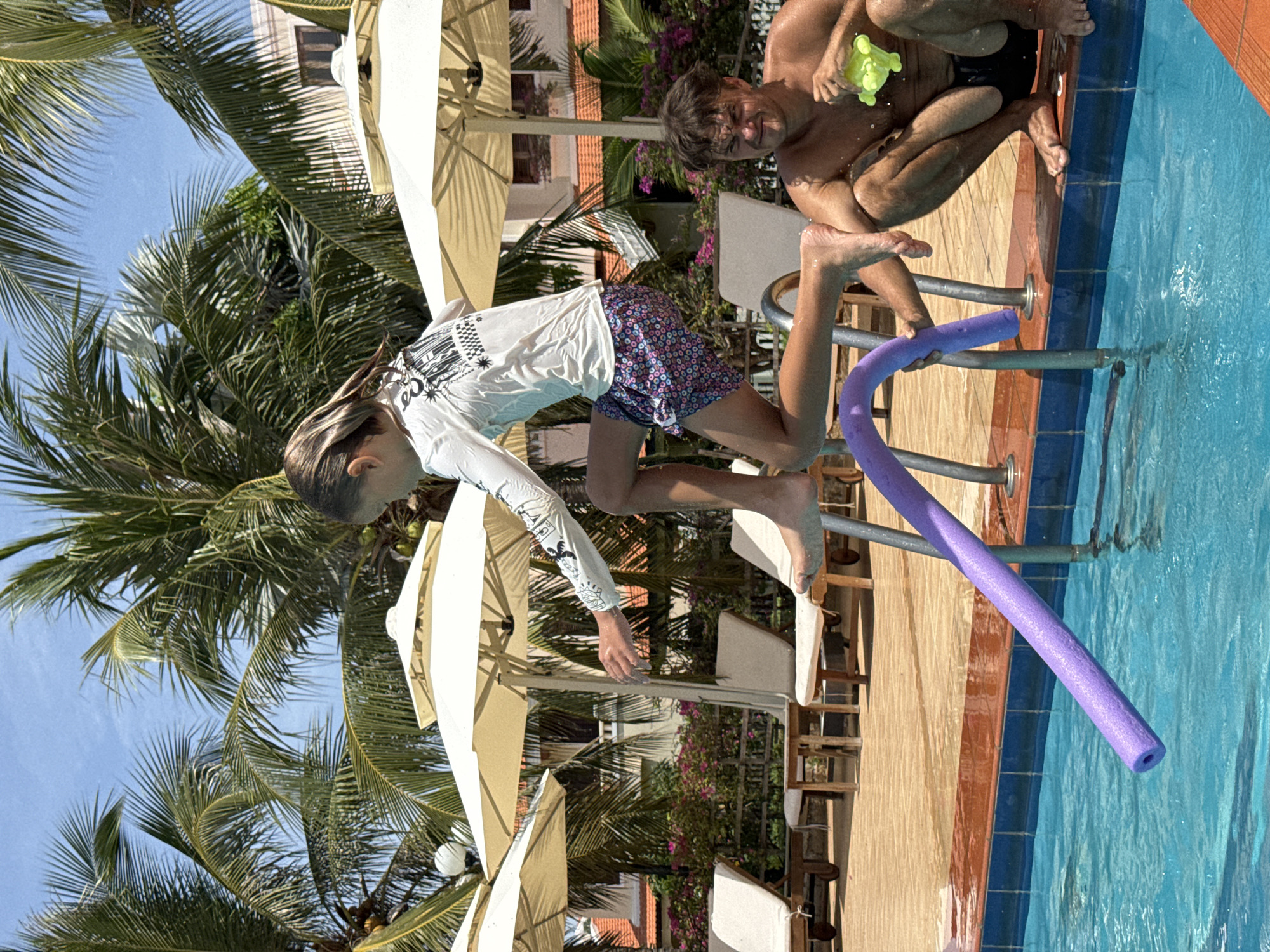 A sunlit, outdoor scene shows a girl jumping into a swimming pool at Phu Thuy Ward, Vietnam. The pool is bright blue, with a purple foam noodle nearby. An adult male sits on the pool edge holding a yellow water toy. The background features palm trees, white umbrellas, and building exteriors. The girl is wearing a white long-sleeved shirt and patterned swim trunks. The man is shirtless.