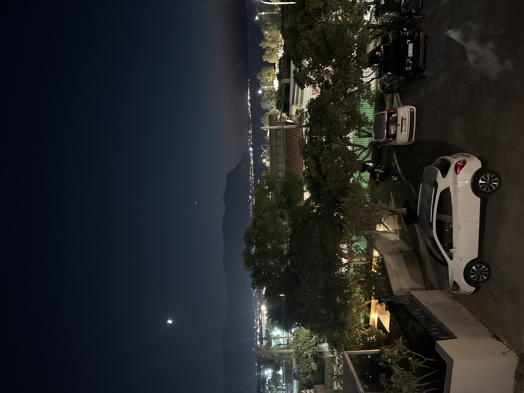 The image depicts a nighttime view of the city of Capaci, Italy, from an elevated perspective. In the dark sky, a crescent moon is visible, along with a few stars. Below the sky, the distant cityscape is illuminated by numerous lights, indicating a populated area. A body of water, likely the sea, stretches across the middle ground, with lights from boats or coastal structures reflecting on its surface.
In the foreground, a parking lot is visible with several parked vehicles. A white SUV is parked on the left, while a white hatchback, a black sedan, and a motorcycle are parked further back. Trees and foliage are scattered throughout the scene, some illuminated by artificial lights. The overall impression is of a peaceful night in a coastal Italian town.