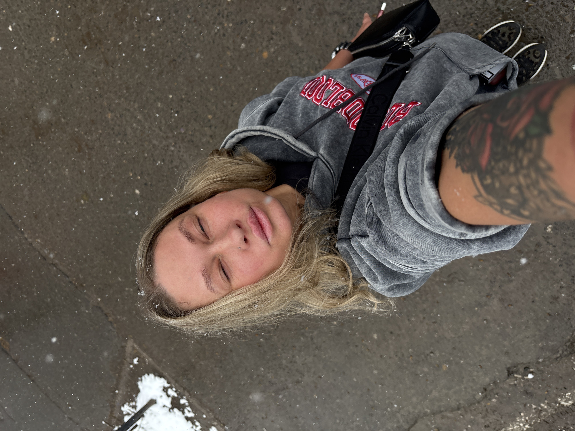 A first-person perspective photograph, taken outdoors during snowfall, shows a woman in a gray hoodie with red lettering, black bag, and black shoes. The location appears to be a city street with a concrete surface covered with snow. A portion of the woman's tattooed arm is visible. The woman's eyes are closed, and she appears to be looking upwards. The bag is slung across her body. Based on the red lettering on the hoodie, and the request in the prompt, the location is potentially Perm, Russia, though this is not explicitly confirmed in the image.