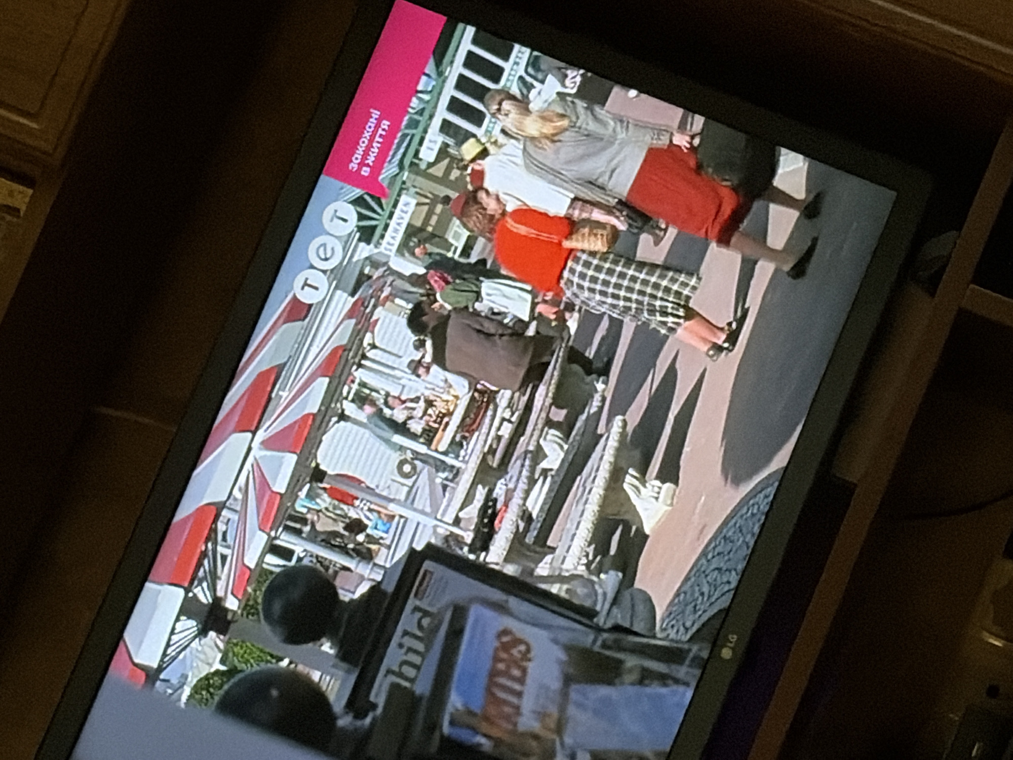 The image shows a television screen displaying a scene of people in what appears to be an outdoor market or fair. There are several people visible, some walking and some sitting. In the foreground, there is a television screen displaying an image of a scene with people under a red and white striped canopy. The TV screen has a logo that appears to be "LG". There is also text on the TV screen that reads "SEAHAVEN" and in a pink box "закохані в життя" which translates to "in love with life". The image is taken at an angle, suggesting it is a photograph of a television screen. The location in the city of Osolynka, Ukraine, is not depicted in this image.