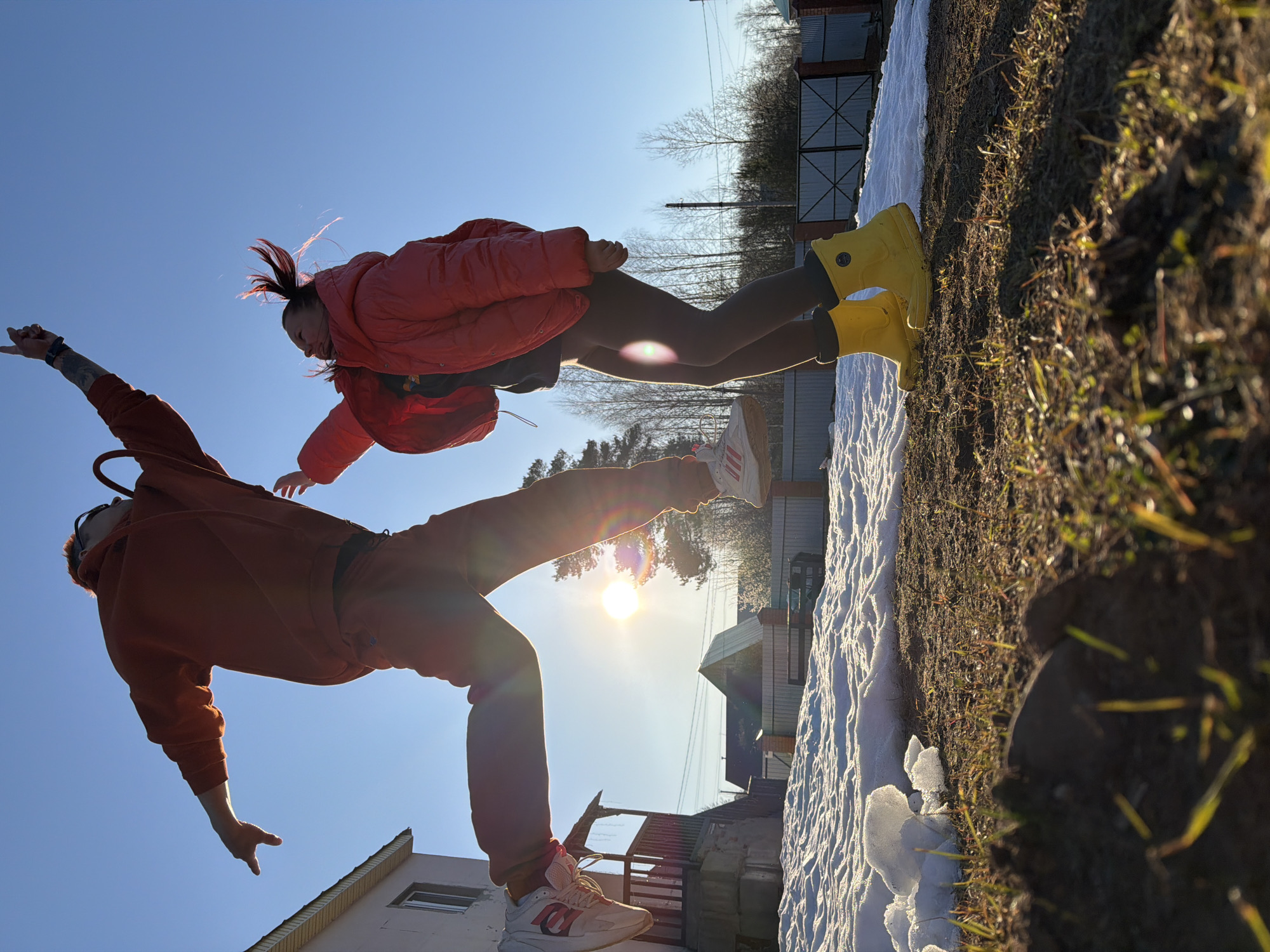 The image captures two individuals in motion against a blue sky, possibly in Chaikovsky, Russia. A person in an orange hooded sweatshirt and matching pants is airborne, with one leg extended towards the sun. The person wears white and orange sneakers. Beside them, another person in a pink jacket, dark leggings, and yellow rain boots is also suspended in the air. Below, there is a landscape transitioning from snow-covered ground to grass. Buildings are visible in the background. The scene is brightly lit by the sun.