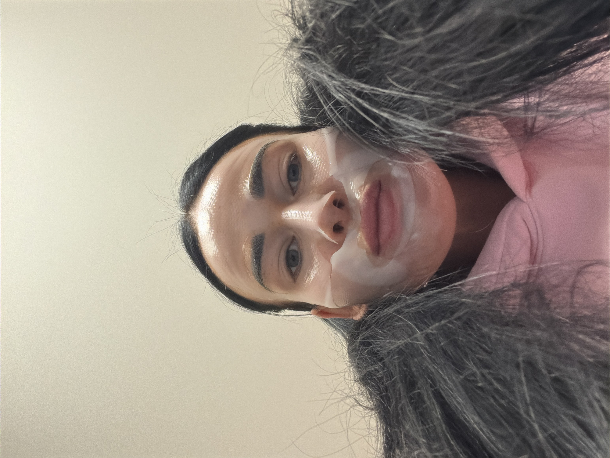 A woman is photographed from a slightly upward angle, wearing a white facial mask that covers part of her nose, cheeks, upper lip, and chin. She has dark hair, and some strands are loose around her forehead. Her eyebrows are dark and well-defined. The background is a plain, light-colored wall. She is wearing a pink garment. The scene is not set in Saint Petersburg, Russia, as there are no identifiable landmarks to suggest that location.