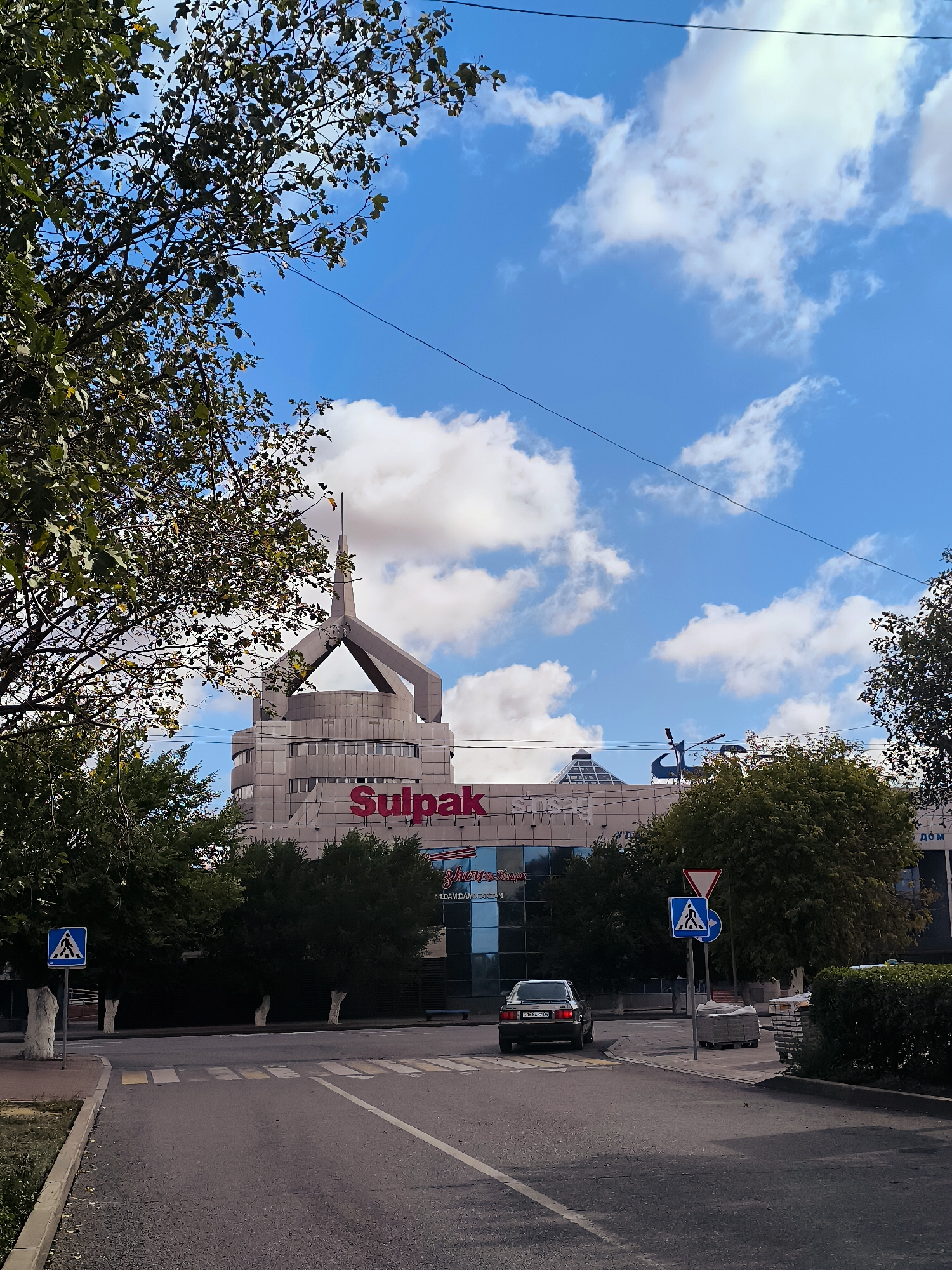The image shows a street view in Qarağandı, Kazakhstan, with a prominent building in the background. The building, identified as the Qarağandı city administration building, features a modern architectural design with a distinctive peaked roof structure. The facade is light-colored, and large red lettering on the building reads "Sulpak" and "Sinsay". In front of the building, a row of dark green trees lines the street. The foreground depicts a road with clear lane markings and a crosswalk. A black sedan is visible driving on the road. Two traffic signs are present on the right side of the road: one a yield sign and the other a pedestrian crossing sign. On the left side of the road, another pedestrian crossing sign is visible. The sky above is blue with scattered white clouds. Trees with green and some yellowing leaves are visible in the left portion of the image, partially obscuring the sky and the upper part of the building. The overall scene depicts a daytime urban environment.