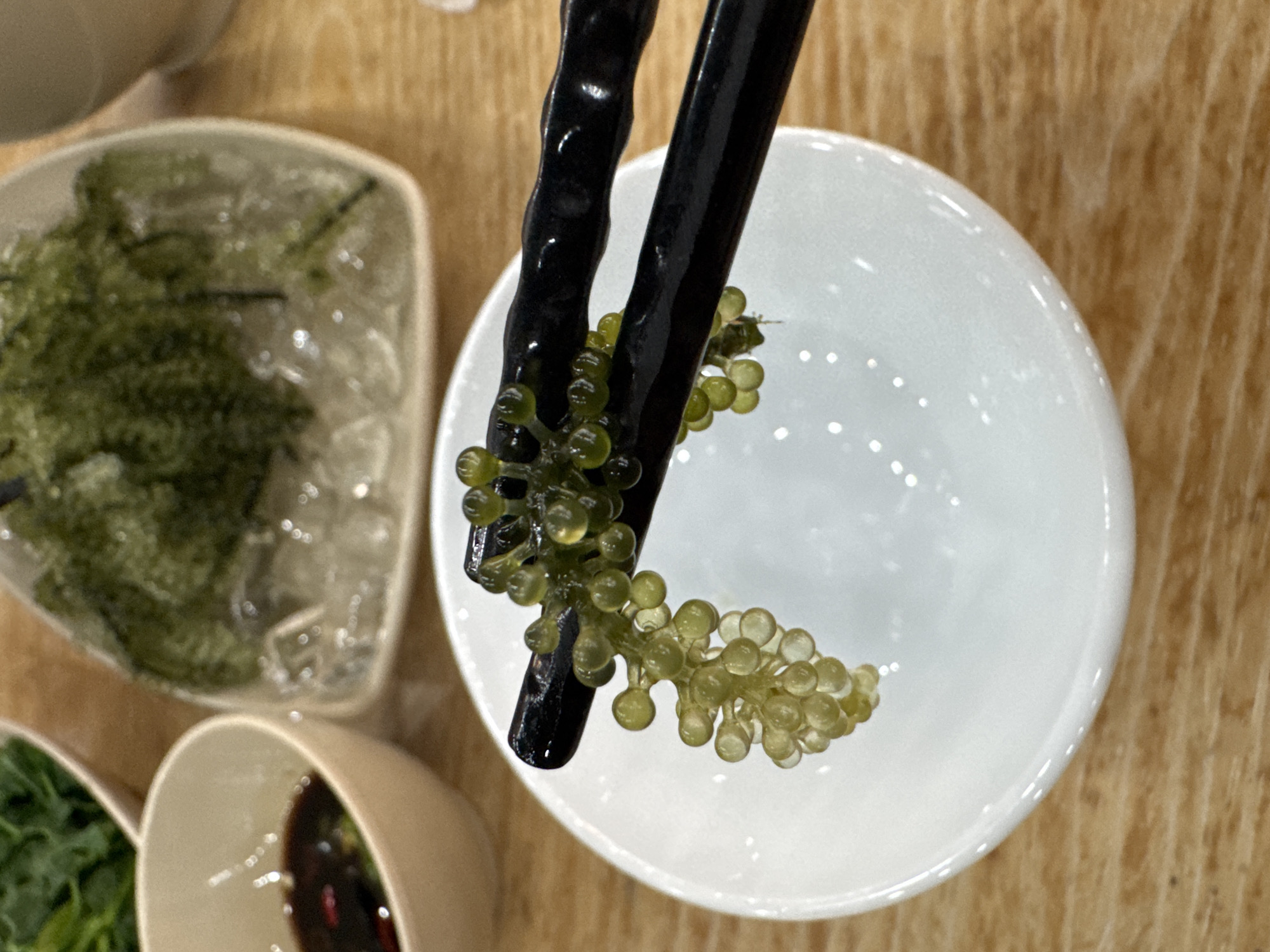 The image depicts a pair of black chopsticks holding a cluster of sea grapes (caulerpa lentillifera) over a small, empty white bowl. In the background, there are other bowls with what appears to be more sea grapes and dipping sauce. The scene likely takes place in a restaurant in Đà Nẵng, Vietnam, as sea grapes are a local delicacy.