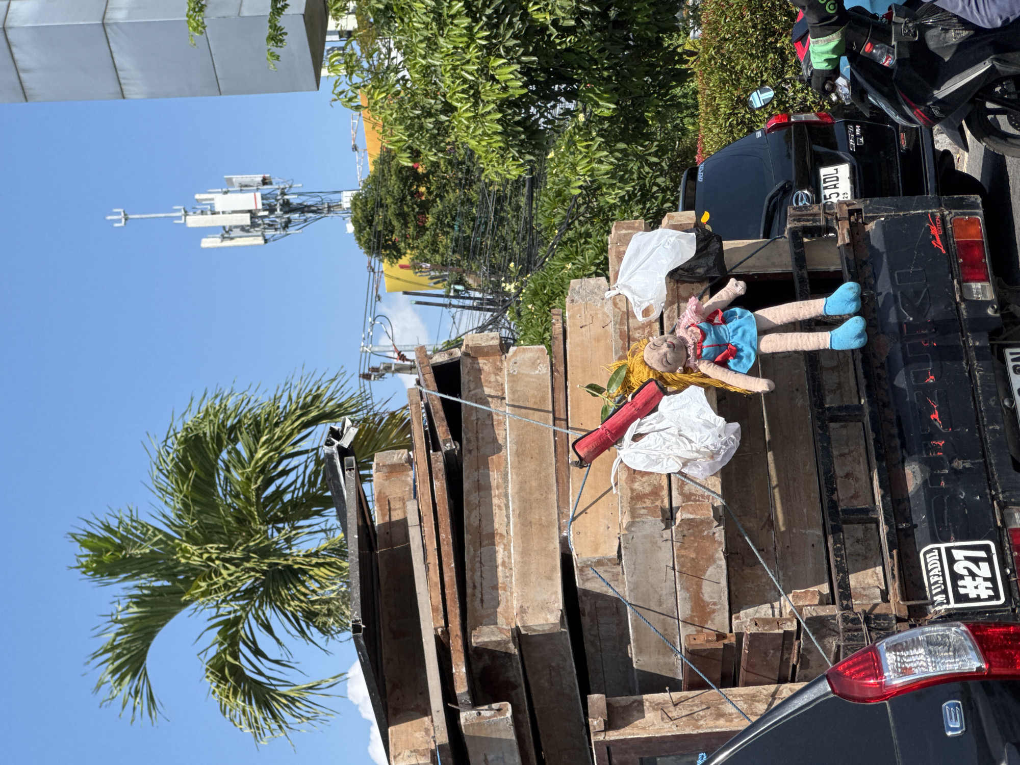 In Kuta, Indonesia, a flatbed truck is laden with a stack of weathered wooden boards, possibly for construction or renovation. A large, handmade doll with blonde hair, a blue dress, and pink trim is propped against the wood, its feet clad in blue. Two white plastic bags are also secured to the load. Behind the truck, a black SUV is parked, and a motorcycle with a rider wearing a green and black jacket is visible on the right. The scene is set against a backdrop of lush green trees and a clear blue sky, with a telecommunications tower and a modern building in the distance. The truck itself is black and displays a license plate with the number "#21" and the name "MU.FADIL".