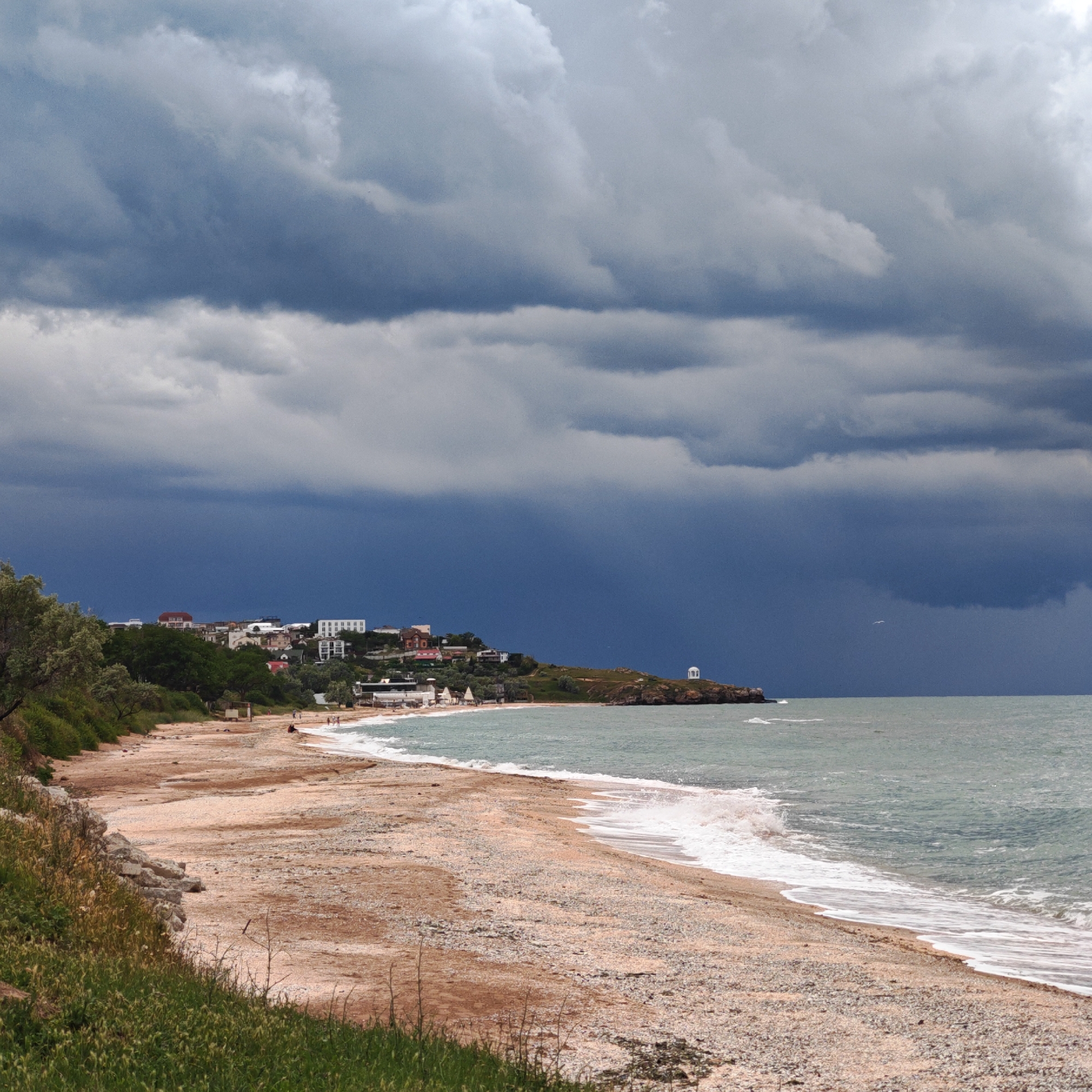 The image captures a coastal scene in городское поселение Щелкино, Ukraine under a dramatic sky. A wide, sandy beach extends into the frame, merging with a shoreline composed of small pebbles. Gentle waves lap against the shore, creating a foamy edge. To the left, grassy vegetation covers a slight embankment. A cluster of buildings, predominantly residential, is nestled along the elevated terrain in the distance. A small, white gazebo stands prominently on a rocky promontory extending into the sea on the right. The sky is dominated by heavy, dark storm clouds, contrasting with the lighter tones of the beach and sea. A single bird is visible in the sky. People are faintly visible on the beach in the distance.