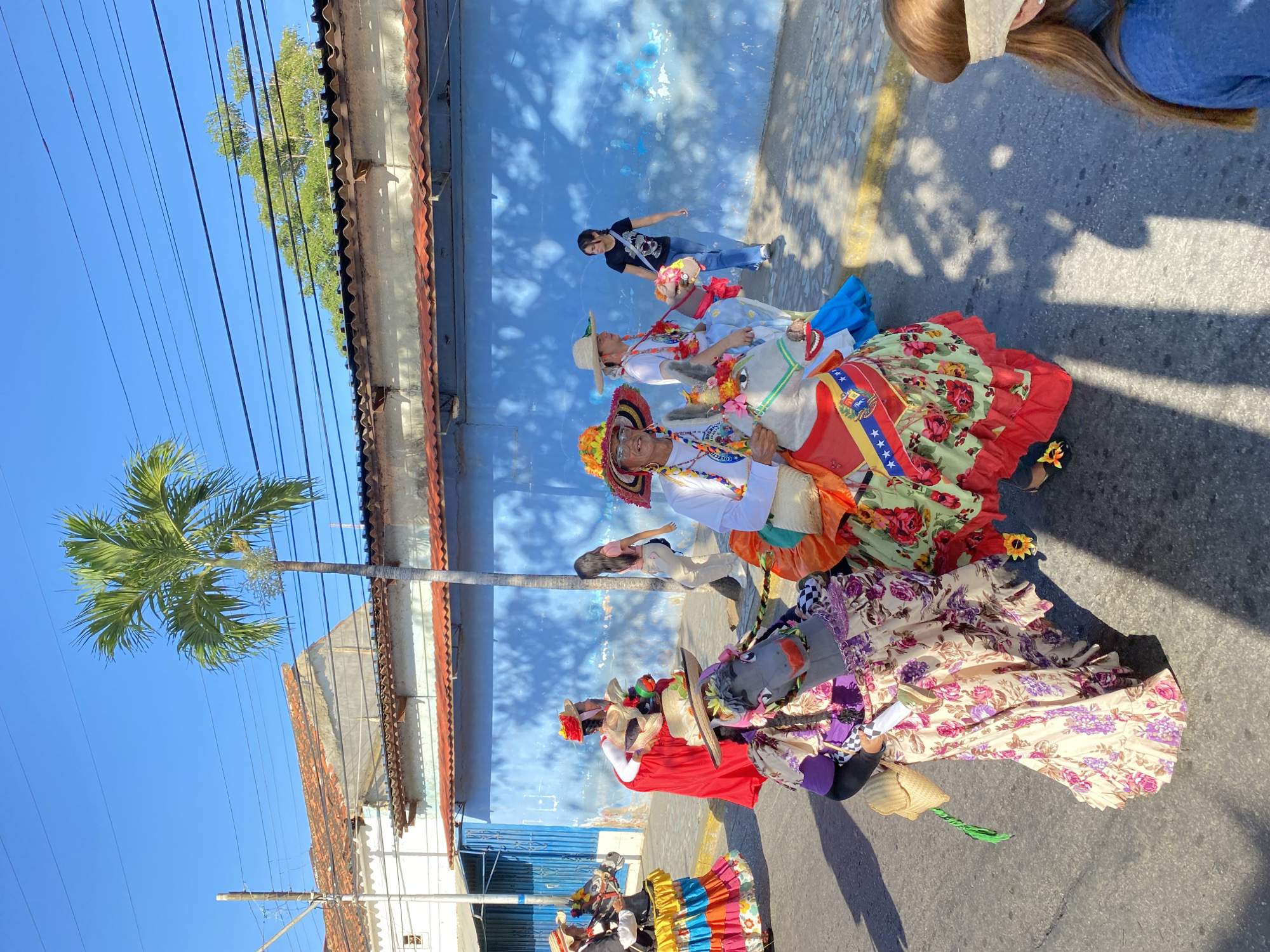 The image depicts a street scene in Juan Griego, Venezuela. The photograph appears to be taken at ground level during the day. Several individuals are riding or walking alongside donkey-like figures. These are either real donkeys or people dressed as donkeys. The individuals are wearing vibrant, folkloric costumes and straw hats adorned with flowers. One donkey-like figure is covered with floral fabric. One of the dresses appears to be a red, green and floral design. The dresses vary in color. The donkey figures have colorful sashes. One sash features the Venezuelan flag. In the background, a blue building with a textured wall is visible. A palm tree is visible on the left side of the frame. Overhead, there are electrical wires. The sky is clear and blue. Shadows indicate a sunny day. A couple of people are walking on the edge of the shot. A woman in the top right corner is wearing a hat and mask.