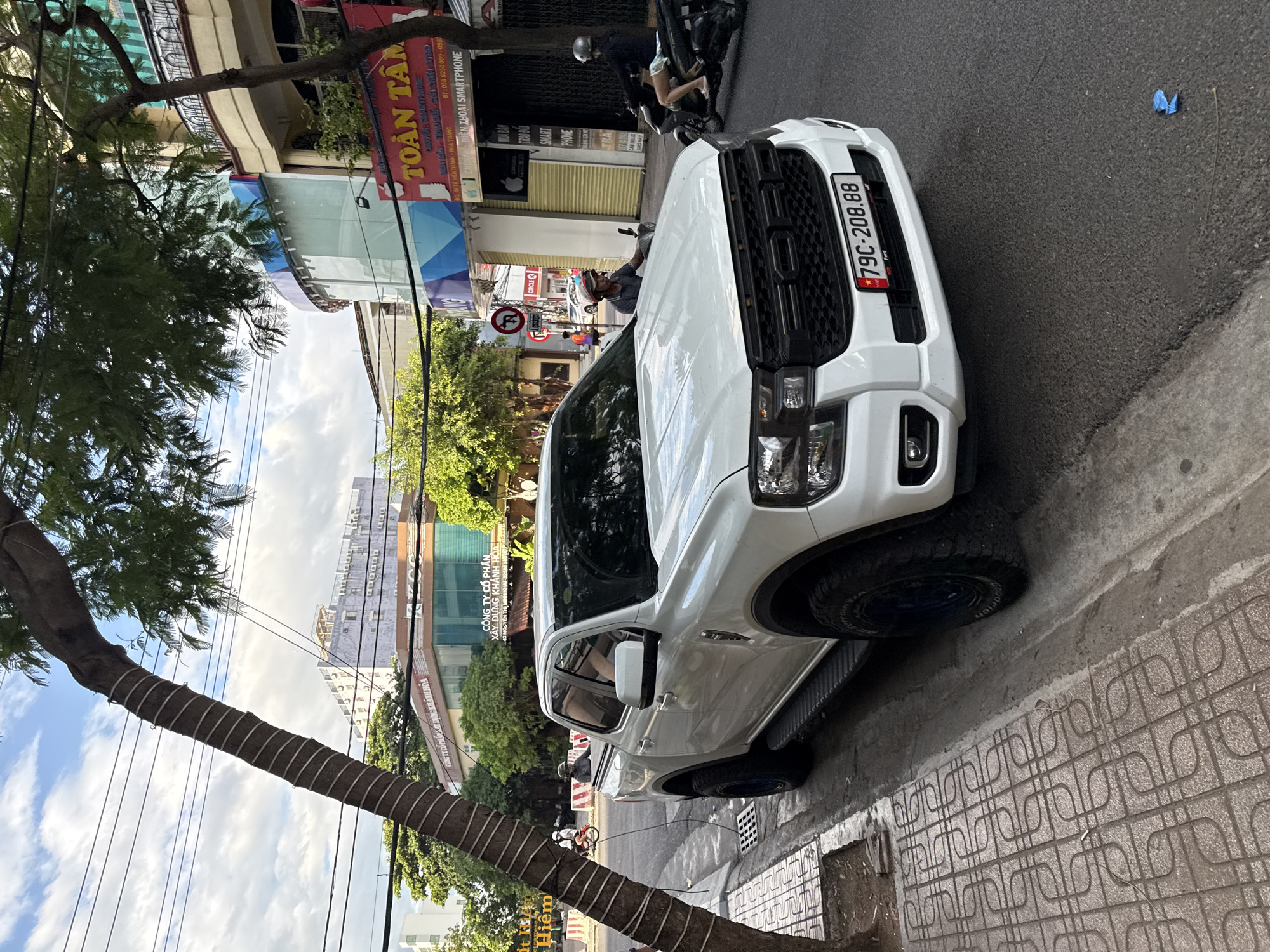 A white Ford Ranger pickup truck is parked on the side of a street in Nha Trang, Vietnam. The truck has a black grille and tinted windows. The license plate reads "79C-208.88". The truck is parked on the right side of the road, adjacent to a sidewalk made of patterned tiles. A tree with a thick trunk and leafy branches overhangs the street, with power lines running through its branches. In the background, there are several buildings, including a modern building with a glass facade and a sign that reads "TOÀN TÂM". There are also several motorbikes and people on the street, some of whom appear to be wearing helmets. The sky is partly cloudy.