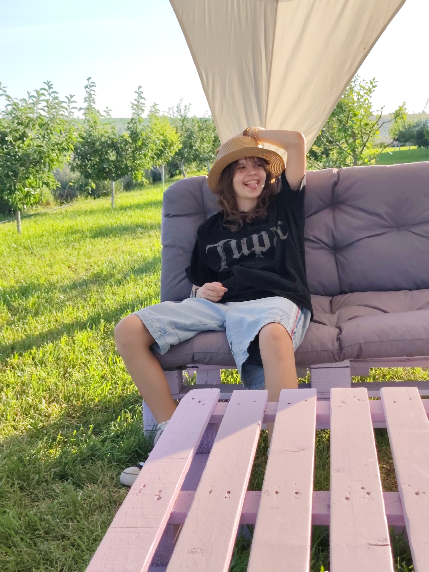 In Nechaiane, Ukraine, a young person wearing a straw hat, a black t-shirt, and denim shorts is sitting on a cushioned sofa outdoors. The sofa is made from stacked wooden pallets painted pink and purple. The person is leaning back on the sofa, with their left arm raised and bent behind their head, and their tongue is sticking out playfully. The scene is set in a grassy area with rows of young trees, likely an orchard, under a clear sky with a large white canopy overhead. The lighting suggests it is a sunny day.