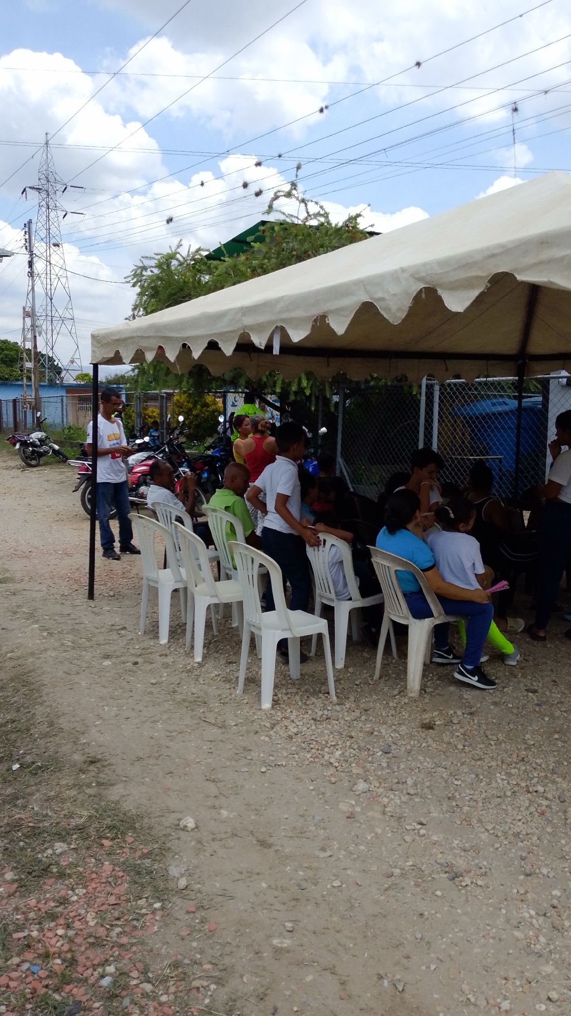 This image depicts an outdoor gathering taking place under a large, light-colored canopy tent in Tinaquillo, Venezuela. The setting features a dirt and gravel ground with scattered small stones, and some sparse grass in the far left foreground.
Underneath the tent, a group of people, comprising both adults and children, are seated on white plastic chairs. They are dressed casually, primarily in t-shirts and jeans. Several individuals appear to be conversing, suggesting an informal social event or community meeting. Some children are standing around the chairs, observing the activities.
To the left of the tent, a man wearing a white t-shirt, blue jeans, and glasses stands near a tent pole, looking towards the seated group. Behind him, several motorcycles are parked. The background features a chain-link fence. Beyond the fence, green trees and a building with a dark green roof are visible. A prominent metal electricity transmission tower stands in the upper left background, with power lines stretching across a bright, partly cloudy sky. Small, dark, spiky objects are suspended from these power lines at regular intervals.
The scene is illuminated by natural daylight, indicating it is daytime, with scattered white clouds in the sky. The atmosphere appears relaxed and communal. There is no visible text on any of the subjects or objects in the image.