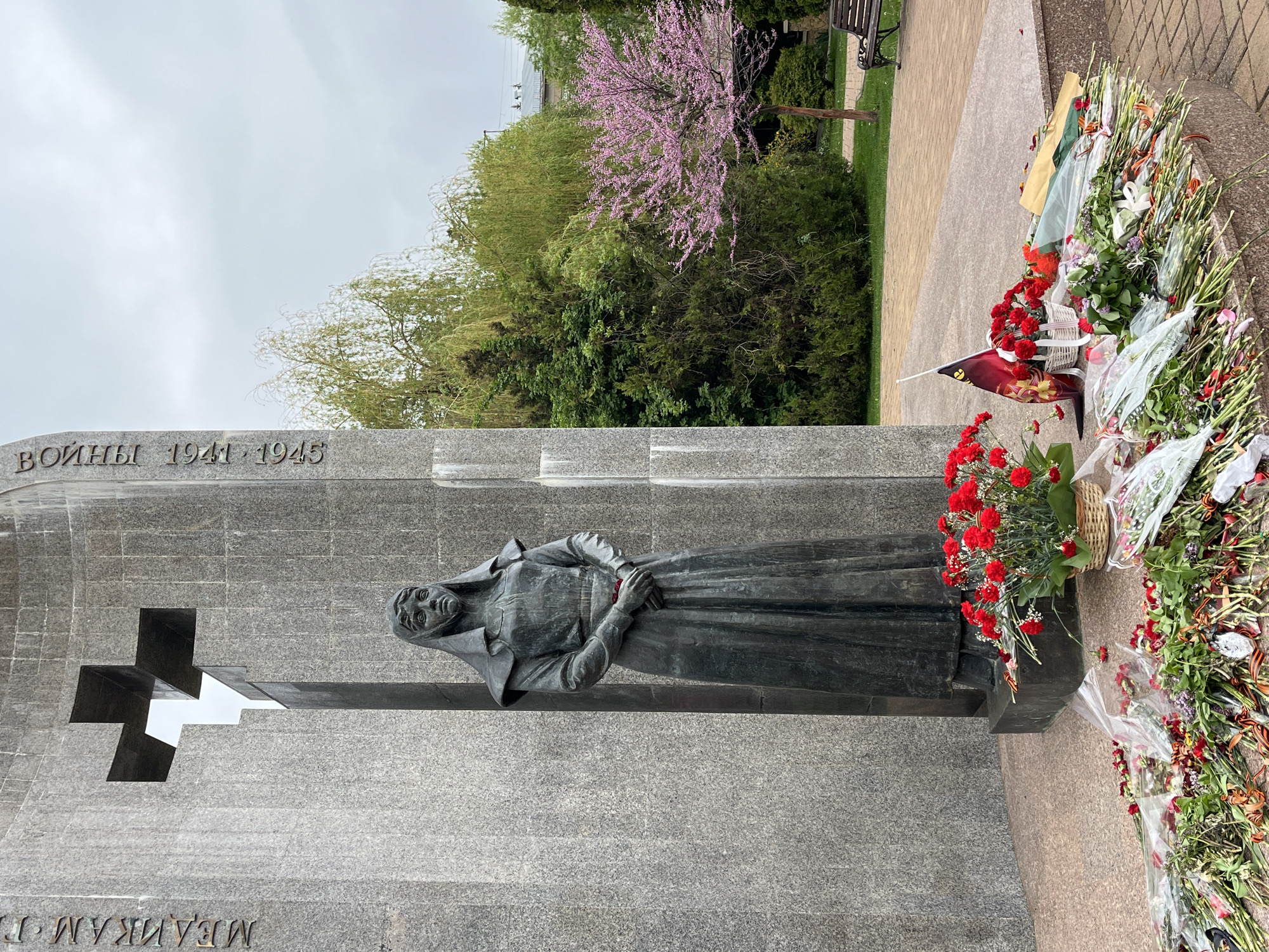 The image shows a statue in Kislovodsk, Russia, commemorating the victims of World War II. The monument features a gray stone wall with an inscription in Russian, a carved cross, and a bronze statue of a grieving woman. Flowers, including red carnations, and other memorial items are placed at the base of the statue. The surrounding environment includes trees, grass, and a paved walkway, suggesting a park or memorial garden setting.