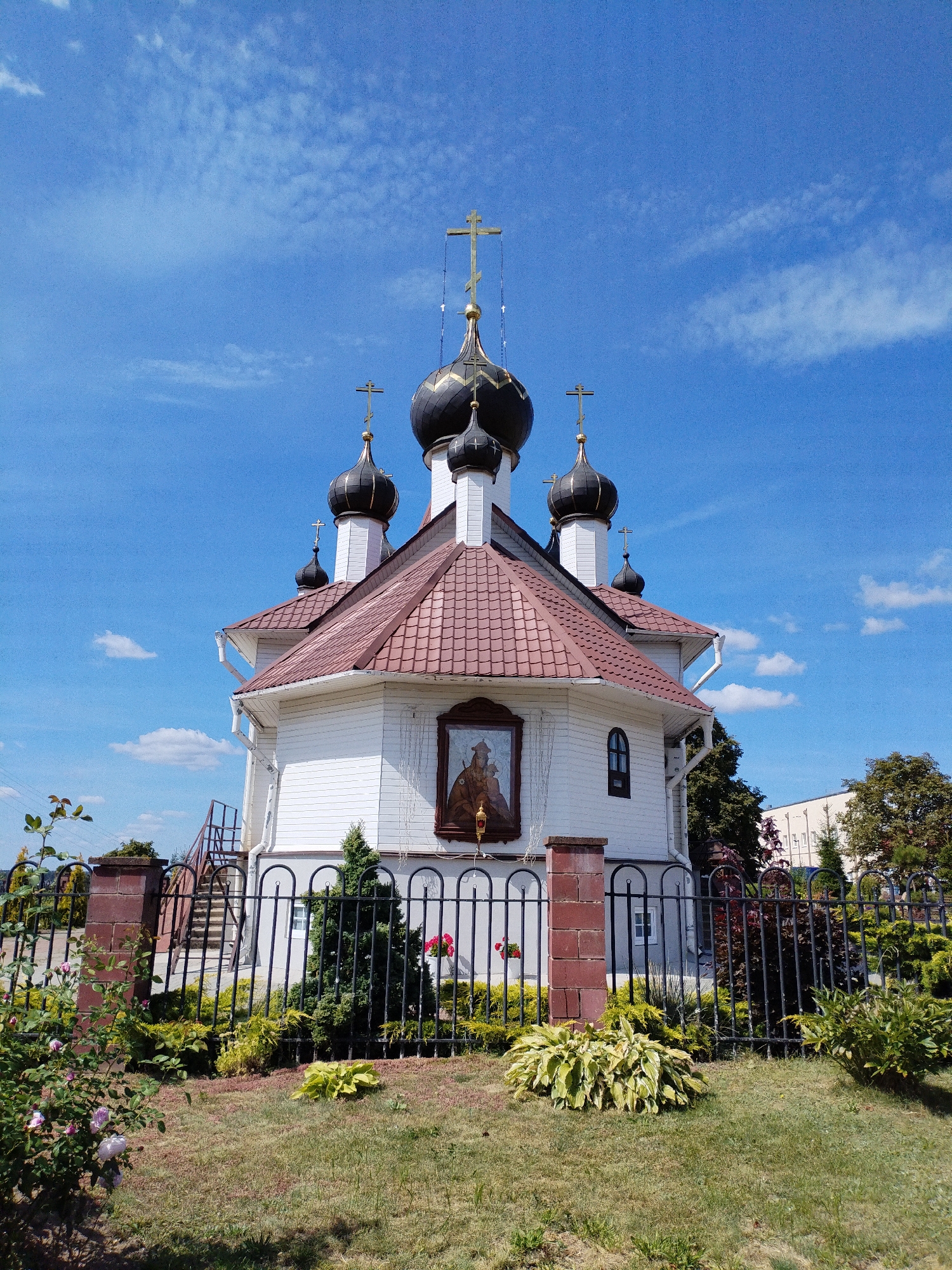 The image shows an Eastern Orthodox church with a white wooden exterior and a reddish-brown tiled roof. The church has multiple black domes topped with golden crosses. A religious icon is mounted on the exterior wall. The church is surrounded by a black metal fence, with a brick pillar supporting one section of it. In front of the fence, there is a garden with various plants, including rose bushes, evergreens, and hostas. The ground is covered with grass. In the background, there are trees and a modern building. The sky is blue with scattered white clouds. This is the Church of the Holy Trinity in the city of Bialynichy, Belarus.