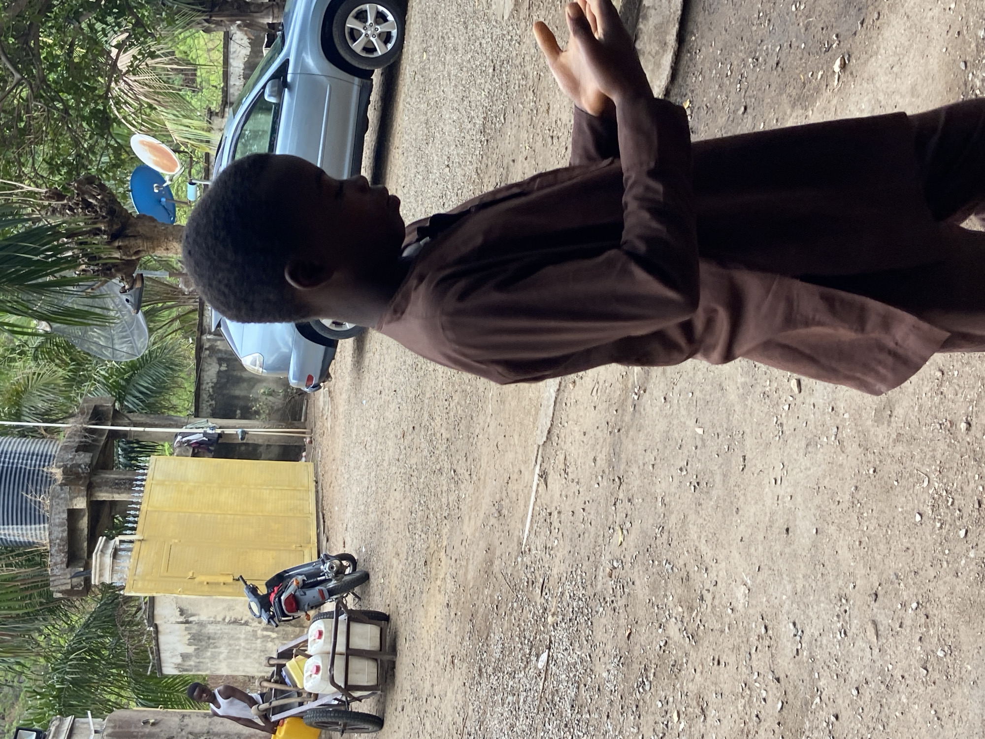 A young boy in a brown shirt is standing in a dirt lot in Mararaba, Nigeria. A silver car is parked nearby. There is a yellow building and a motorcycle attached to a trailer carrying jugs of water. A water tank and trees are in the background.