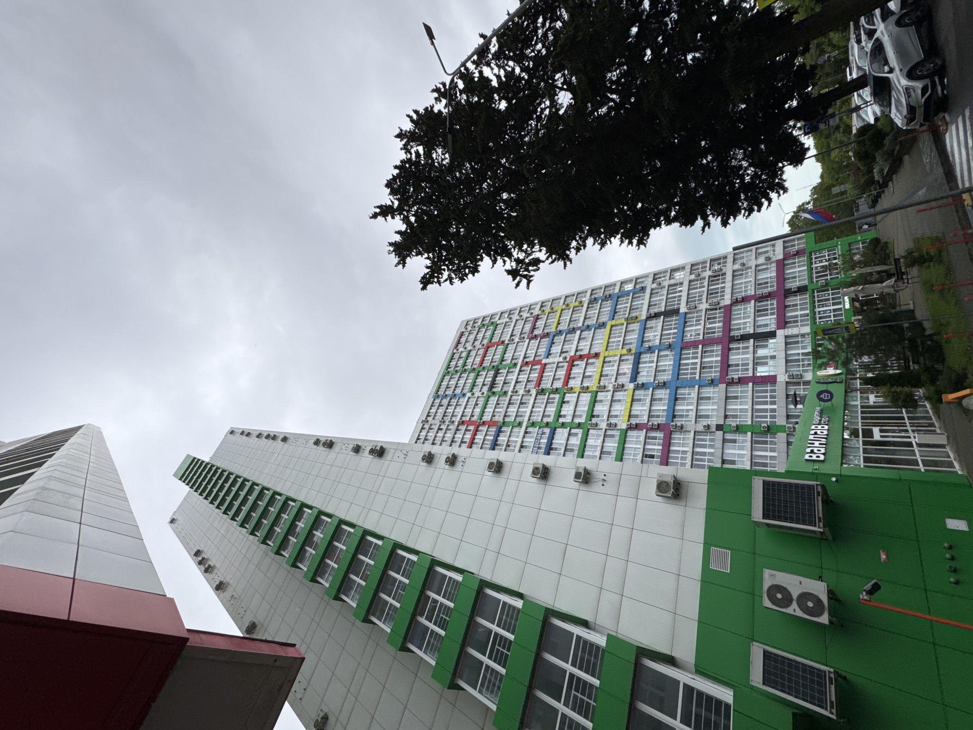 The image shows a low-angle view of several buildings in Sochi, Russia. The sky is overcast. The foreground is dominated by two tall buildings. The building on the left has a white facade with red trim. The building to the right has a white facade with a green accent running vertically, including green window trim. Both buildings have numerous windows.
A taller building is partially visible in the background. This building has a white facade with a multicolored grid pattern (red, green, blue, yellow, purple) resembling the Olympic rings. The building is labeled "Ван Лав" in Cyrillic script.
A tree overhangs the top of the frame, partially obscuring the top portion of the building in the background. A streetlight is visible to the left of the tree.
A street with cars is visible in the upper-right portion of the frame.