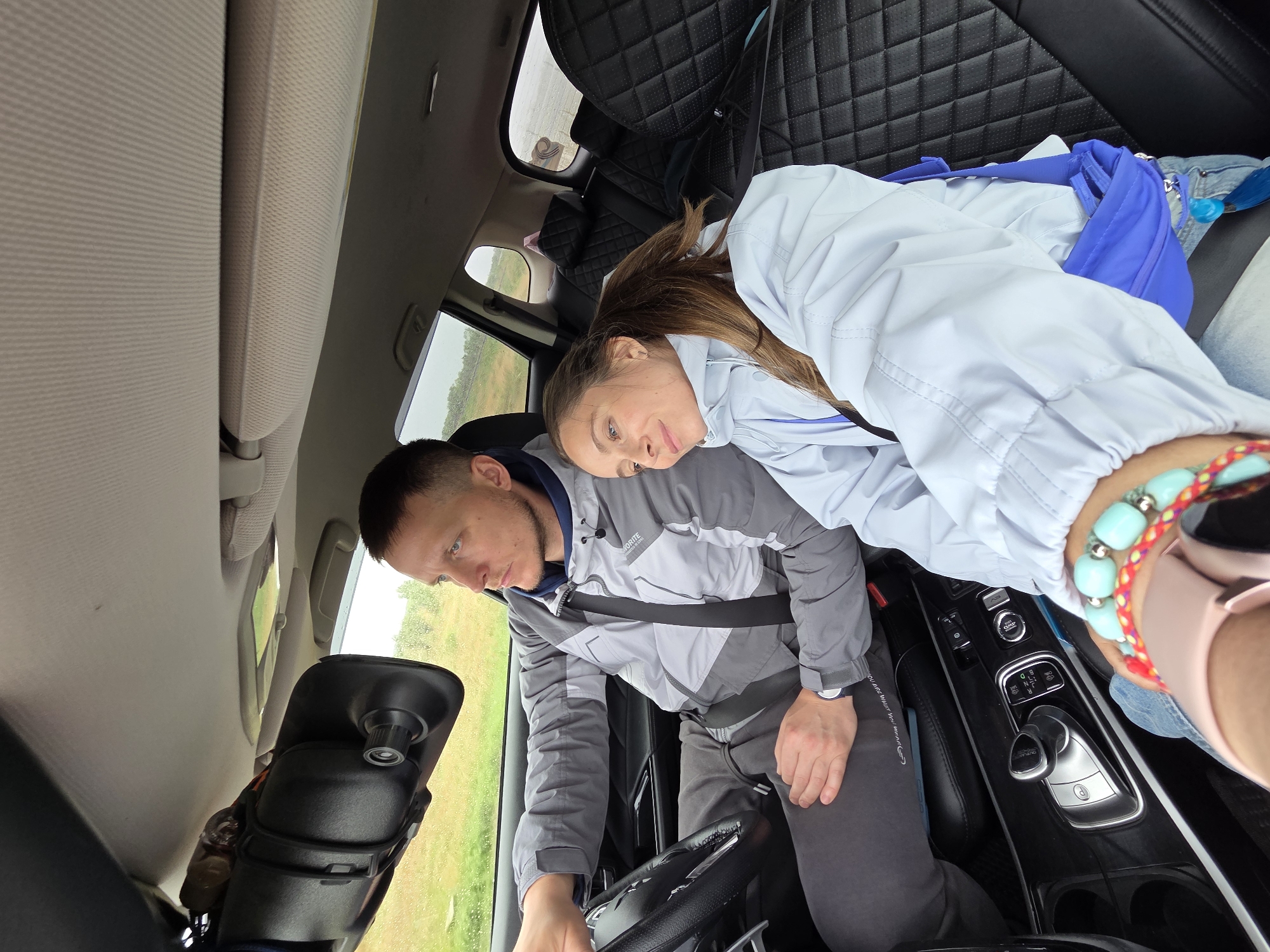 The image captures two people, a man and a woman, inside a car, likely in Бабстово, Russia, during an overcast daytime.
The man is seated in the driver's seat, wearing a two-toned grey jacket with "FAVORITE" and "SPORTSWEAR" visible on it, and a seatbelt across his chest. He looks down intently, possibly at the steering wheel or controls, with a focused expression. A watch is visible on his left wrist.
Leaning against the man's shoulder and looking towards the camera is the woman. She has long brown hair and wears a light blue or light grey jacket. Her expression is soft and gentle. On her right wrist, she wears a multi-colored beaded bracelet, predominantly teal, and a pink smartwatch. A bright blue bag is tucked beside her.
The car's interior features black, diamond-quilted leather seats and a dashboard with an automatic gear shift (P, R, N, D, M settings are visible) and various control buttons. A dashcam is mounted on the rearview mirror. Through the car windows, a green, grassy landscape is visible under what appears to be a cloudy sky, indicating overcast weather.
