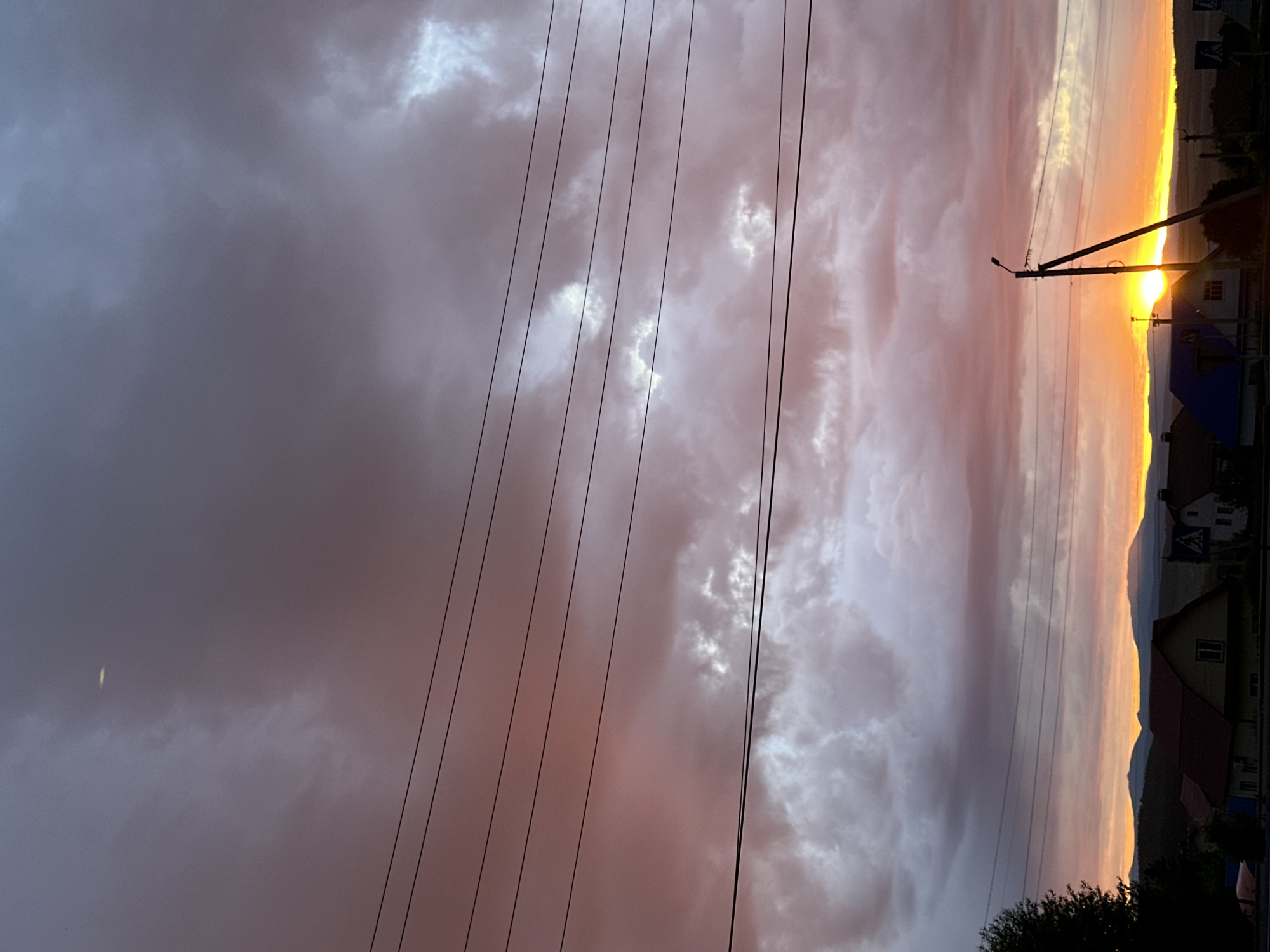 The media file captures a dramatic sunset or sunrise over a semi-rural landscape. The image is dominated by a vibrant and cloudy sky, painted with hues of warm orange, golden yellow, soft pink, purple, and various shades of gray. The sun, a bright, intense orb, is visible low on the horizon towards the right side of the frame, casting a strong glow that illuminates the clouds above and creates a shimmering line on the distant land.
Several dark power lines stretch horizontally across the mid-ground and upper portions of the sky. A prominent wooden utility pole with a street light attached stands on the right, partially silhouetting against the glowing horizon and obscuring a small part of the sun. In the lower section of the image, the silhouettes of distant mountains or hills frame the horizon line. Below these, a collection of houses and buildings with varying roof colors (including dark, red, and blue) are visible, suggesting a small settlement. A patch of dark green foliage, possibly a tree, is present in the bottom right corner. A dark road sign is barely discernible in the far upper right, but no text is legible. The overall scene evokes a sense of tranquility and the beauty of natural light at dawn or dusk.