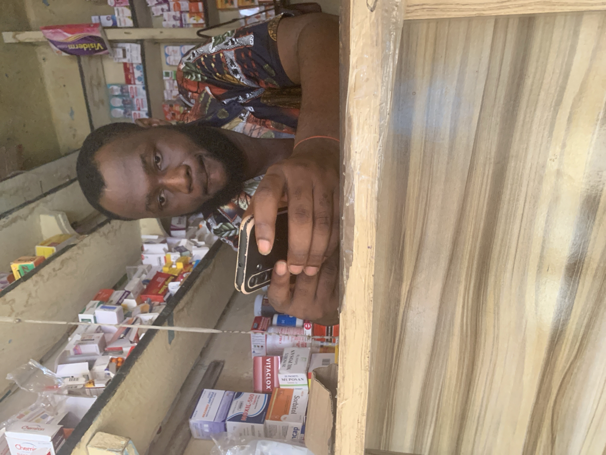 A man with dark skin, short dark hair, and a beard looks directly at the camera. He is wearing a colorful, patterned shirt with hues of red, blue, and orange. His right arm has a thin orange bracelet. He is holding a black smartphone with both hands, positioned in front of him.
The setting is an indoor space, likely a small pharmacy, drug store, or a stall selling medications, indicated by the numerous packaged items on wooden shelves behind him and to his left. The shelves appear somewhat makeshift, filled with various boxes and bottles. A light-colored wooden counter or partition is visible on the right side of the frame, separating the man from the viewer.
The man's expression is neutral to slightly pleasant. He appears to be either posing for the photo or interacting with the photographer.
Visible text on the product packages, aided by OCR, includes "Chemiron" (seen twice), "NOXIL-500", "Visidermo", "VITACLOX", "20 PACKETS MUPOSAN" (seen twice), "Sorbitol", "CALC", "MUPO", and "POSAN". The text "МАЛАМОА САРБИЯ" is also detected, though its legibility is poor. The location context provided is Fegge, Nigeria.