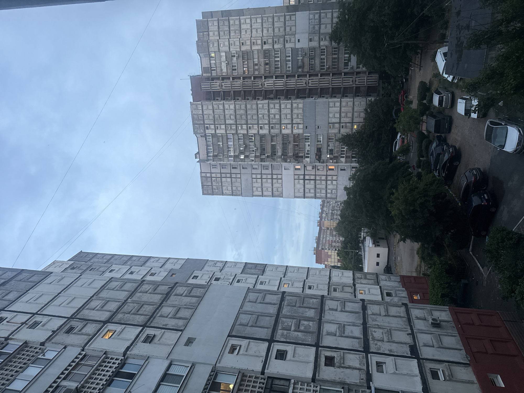 The image depicts a residential area in the city of Dnipro, Ukraine, characterized by Soviet-era apartment buildings. In the foreground to the left, a multi-story apartment building with a grey facade and red lower section is visible. The building features numerous rectangular windows and balconies, with some windows illuminated, suggesting occupancy. To the right, a larger, taller apartment building of similar architectural style dominates the middle ground. This building also has a grey facade and exhibits a grid-like pattern of windows and balconies. The sky above is overcast with a pale blue hue. Below the buildings, a paved courtyard area is visible, containing several parked cars of various models, including sedans, vans, and an SUV. Trees and green foliage are interspersed throughout the courtyard and around the buildings, indicating a somewhat green urban environment. In the distance, more apartment buildings and trees are visible, contributing to the overall urban landscape. The scene appears to be captured during daylight hours, likely in the late afternoon or early evening, given the subdued lighting and illuminated windows.