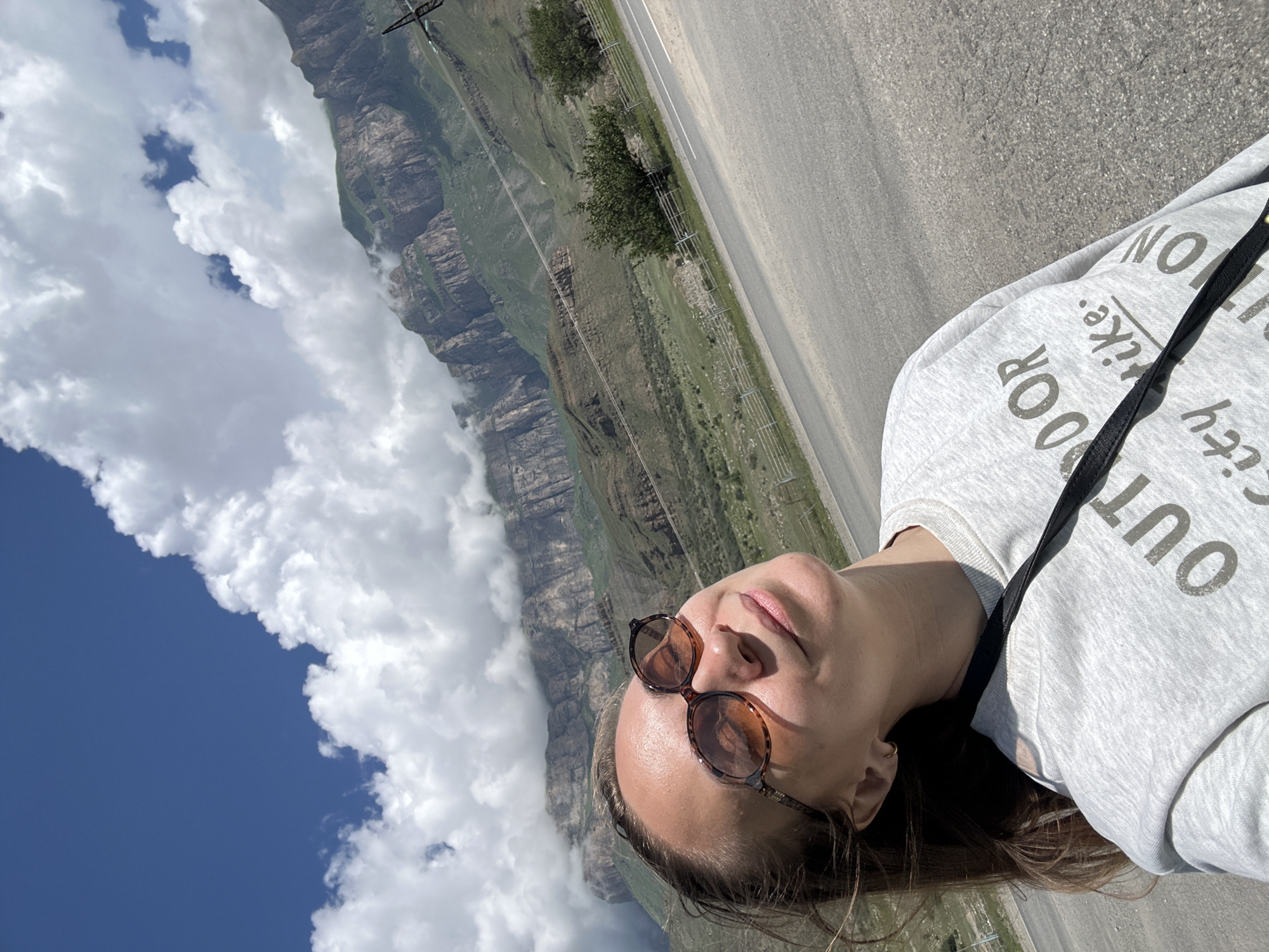 A young woman with brown hair and sunglasses is taking a selfie outdoors. She is wearing a gray sweatshirt with text that appears to read "OUTDOOR CITY LIKE". In the background, there are dramatic mountains with green slopes and rocky cliffs, partially obscured by clouds. A paved road runs in front of the mountains. The location is identified as Bylym, Russia. The weather appears to be sunny with scattered clouds.