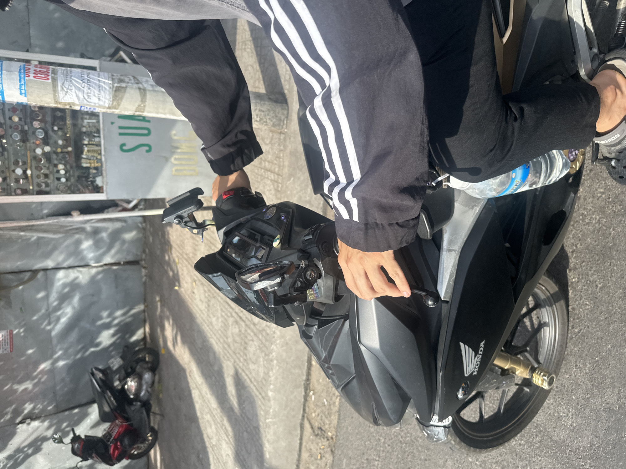 The image depicts a person riding a black Honda motorcycle on a city street. The rider is wearing a black long-sleeved jacket with white stripes on the sleeves and black pants. Their right hand is on the handlebar, and a bottle of water is clipped to the motorcycle. In the background, a red and black motorcycle is parked, and a shop selling watches is visible. The scene appears to be located in Bac Nha Trang Ward, Vietnam, as suggested by Vietnamese text on a pole in the background. The lighting indicates it is daytime, with shadows cast on the ground.