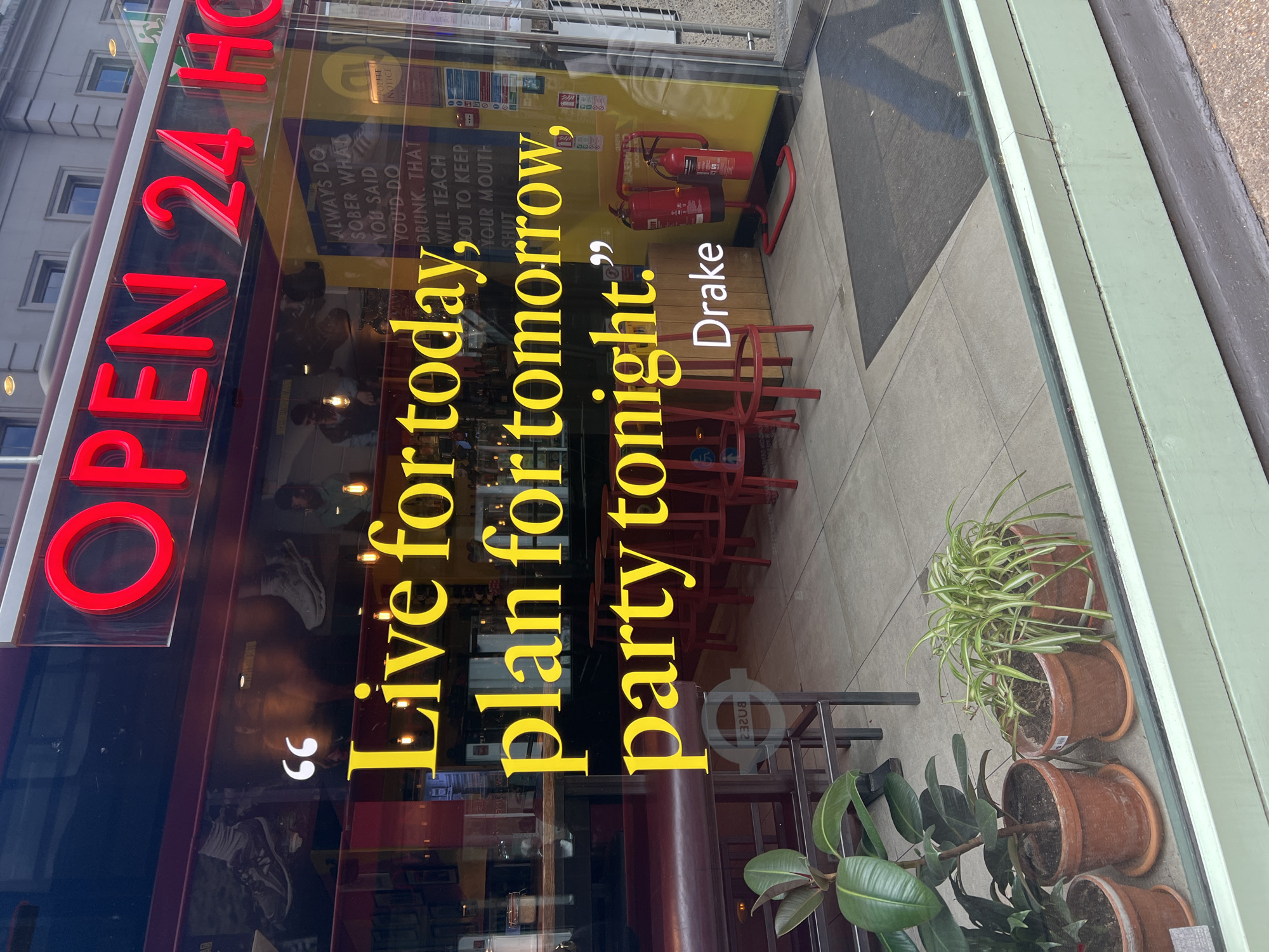 Here's a matter-of-fact description of the image:
The image shows a shopfront in what appears to be London, United Kingdom. The shop is "OPEN 24 H" with the words "OPEN 24" in bright red. The shop window displays a quote attributed to Drake: "Live for today, plan for tomorrow, party tonight." The text is in yellow and vertically aligned on the right. Inside, the shop appears to be a bar or cafe, evidenced by red chairs and a small number of tables. Fire extinguishers are visible on a back wall. A series of potted plants are positioned on the sidewalk outside the shop window. Reflective surfaces show the suggestion of a street and buildings.