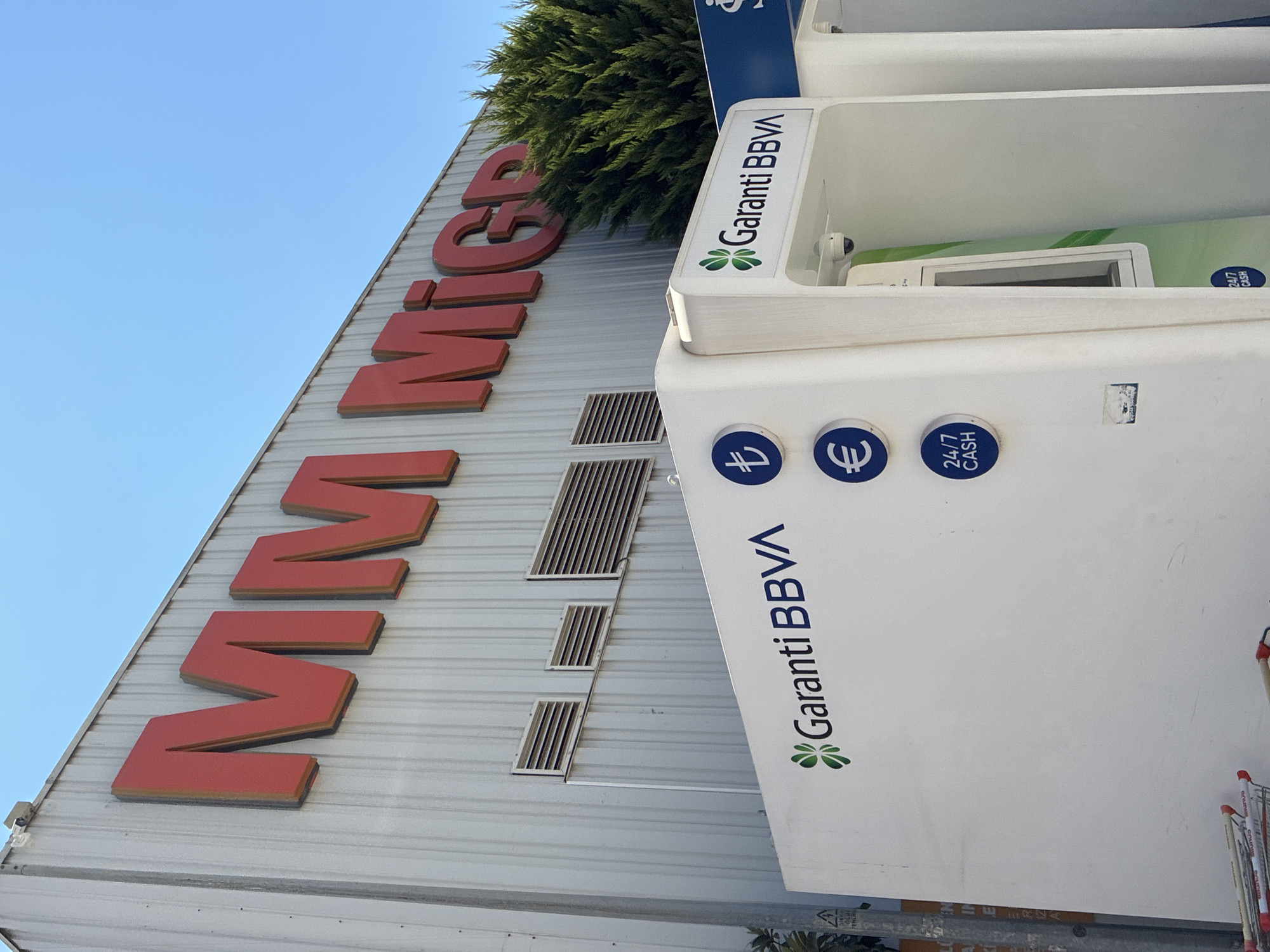 The image shows a close-up of the exterior of a Migros supermarket in Alanya, Turkey. The large red "MM Migros" lettering is prominent on the building's corrugated metal facade. Below the sign, a white kiosk structure is visible, featuring a "Garanti BBVA" logo, indicating a banking service. The kiosk also has circular blue icons for currency symbols (Turkish Lira and Euro) and "24/7 CASH" indicating an ATM. A security camera is mounted on the kiosk. A dense green tree is situated to the right of the building. In the foreground, several shopping carts are partially visible. The sky is a clear blue.
