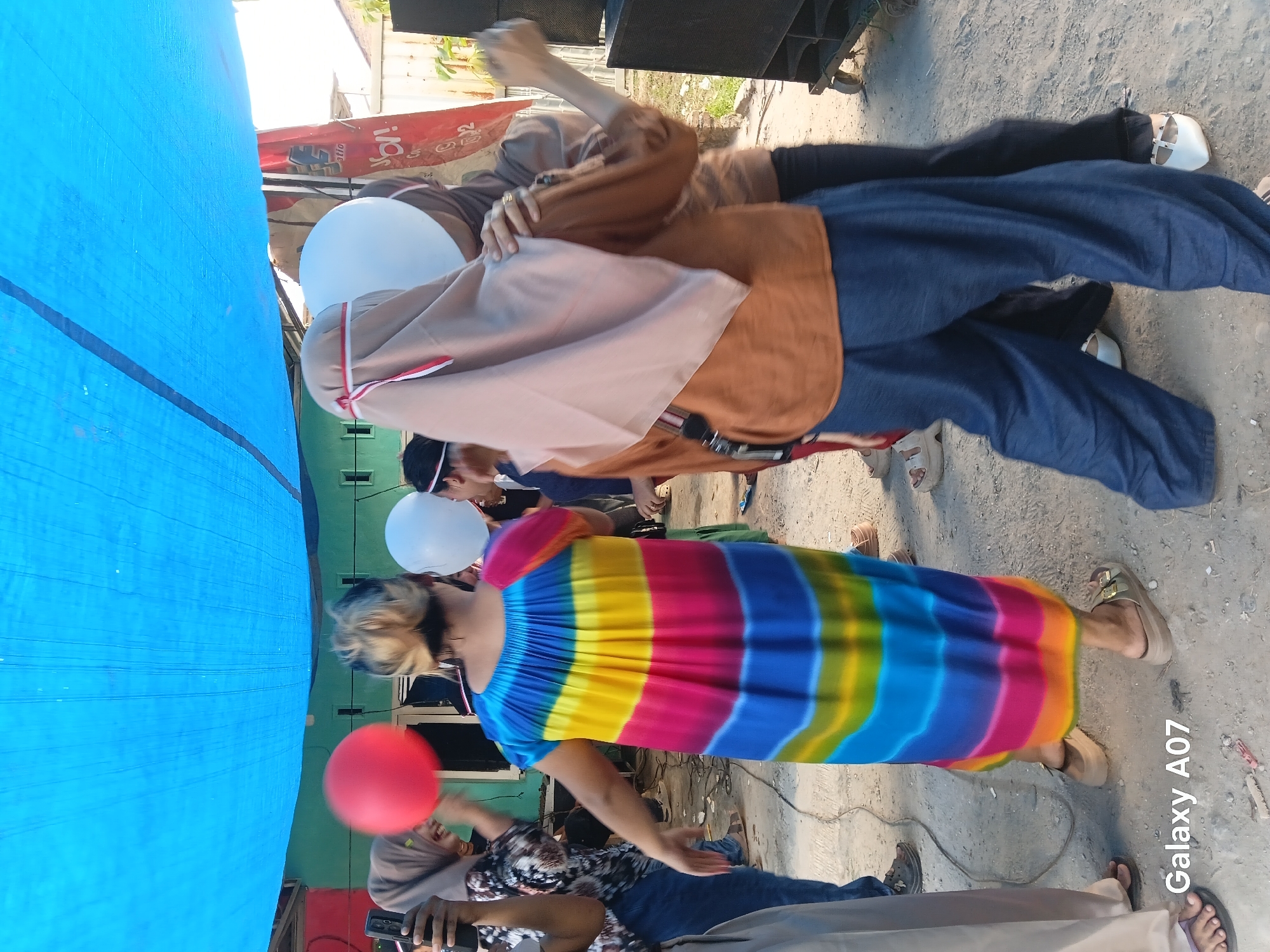 This is a medium shot, eye-level photograph taken outdoors in Tembung, Indonesia. A group of people are gathered under a large blue tarp, possibly for an event or celebration. In the foreground, a person wearing a vibrantly colored, rainbow-striped dress is facing away from the camera, dancing. To their right, another person in a brown shirt and blue pants is also facing away, holding a white balloon. A third person, partially visible, is wearing a brown headscarf and holding a white balloon. To the left, a person in a black and white patterned shirt is holding a red balloon and a mobile phone. The ground is a dirt or gravel surface. In the background, there are buildings and possibly some trees. The lighting suggests it is daytime, with bright sunlight. The overall atmosphere appears to be festive and social.