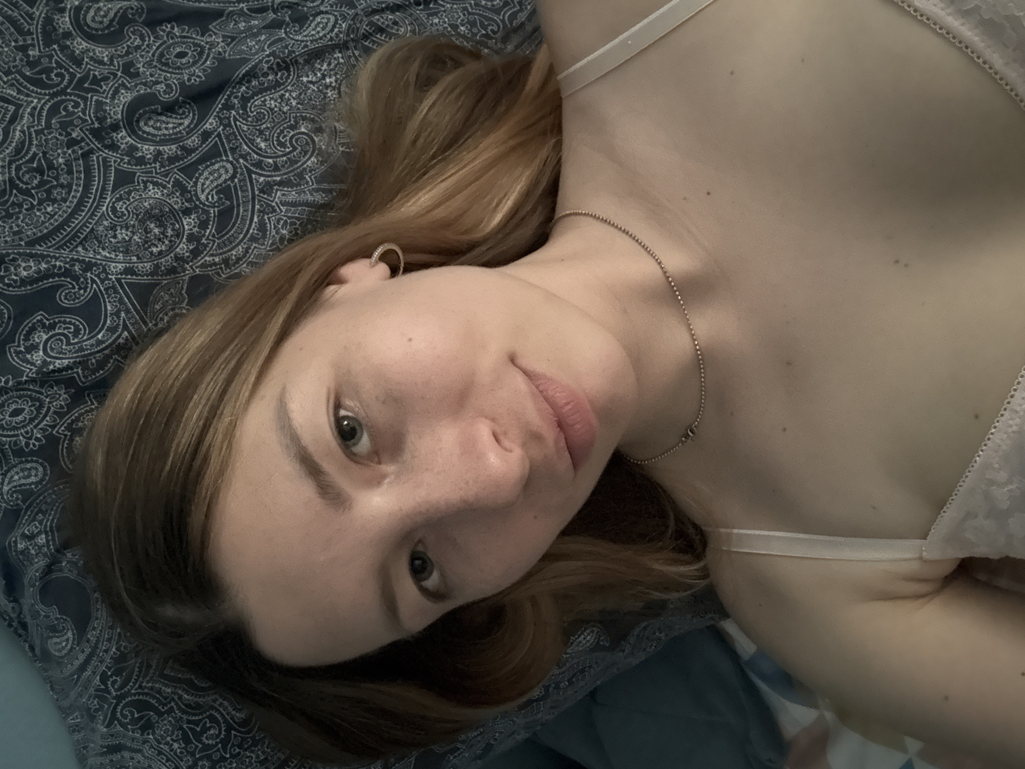 A fair-skinned woman with blonde hair is lying on a bed with a blue paisley patterned sheet. She is wearing a light-colored bra with thin straps and a delicate lace pattern. A thin silver chain necklace adorns her neck. Her face is turned towards the camera, and she has fair skin with freckles. The location is not discernible from the image.